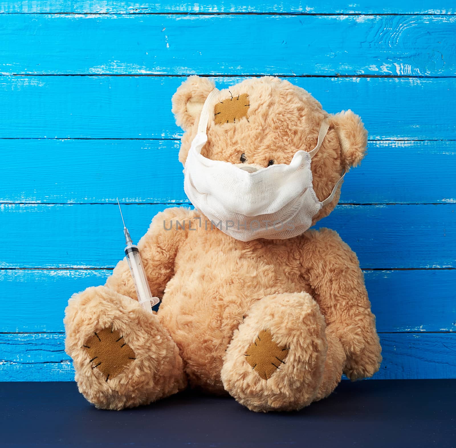 beige big teddy bear with patches sitting in a white medical mask in the paw of a syringe, concept of the need for vaccinations in children, blue wooden background. Injection, vaccine concept.