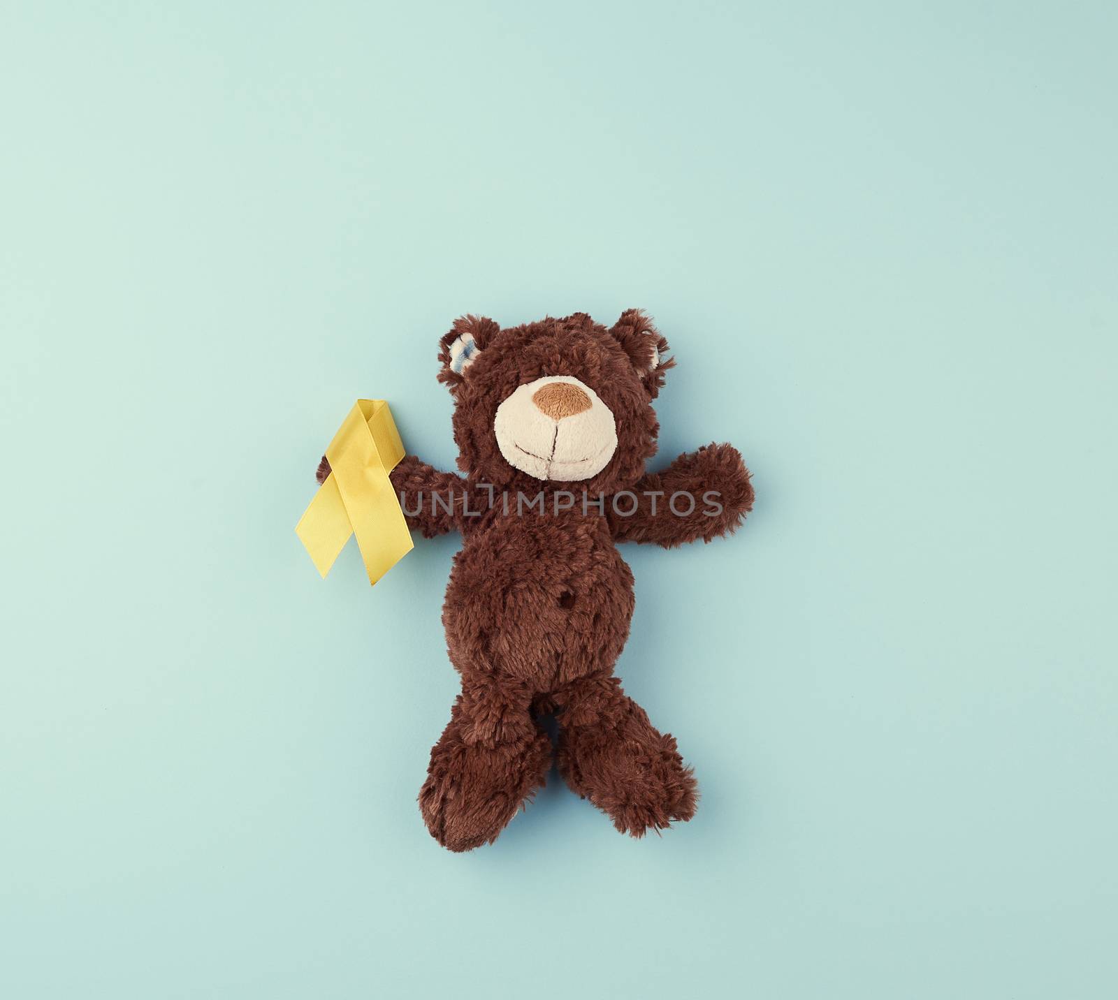 brown teddy bear holds in his paw a yellow ribbon folded in a loop on a blue background. concept of the fight against childhood cancer. problem of suicides and their prevention