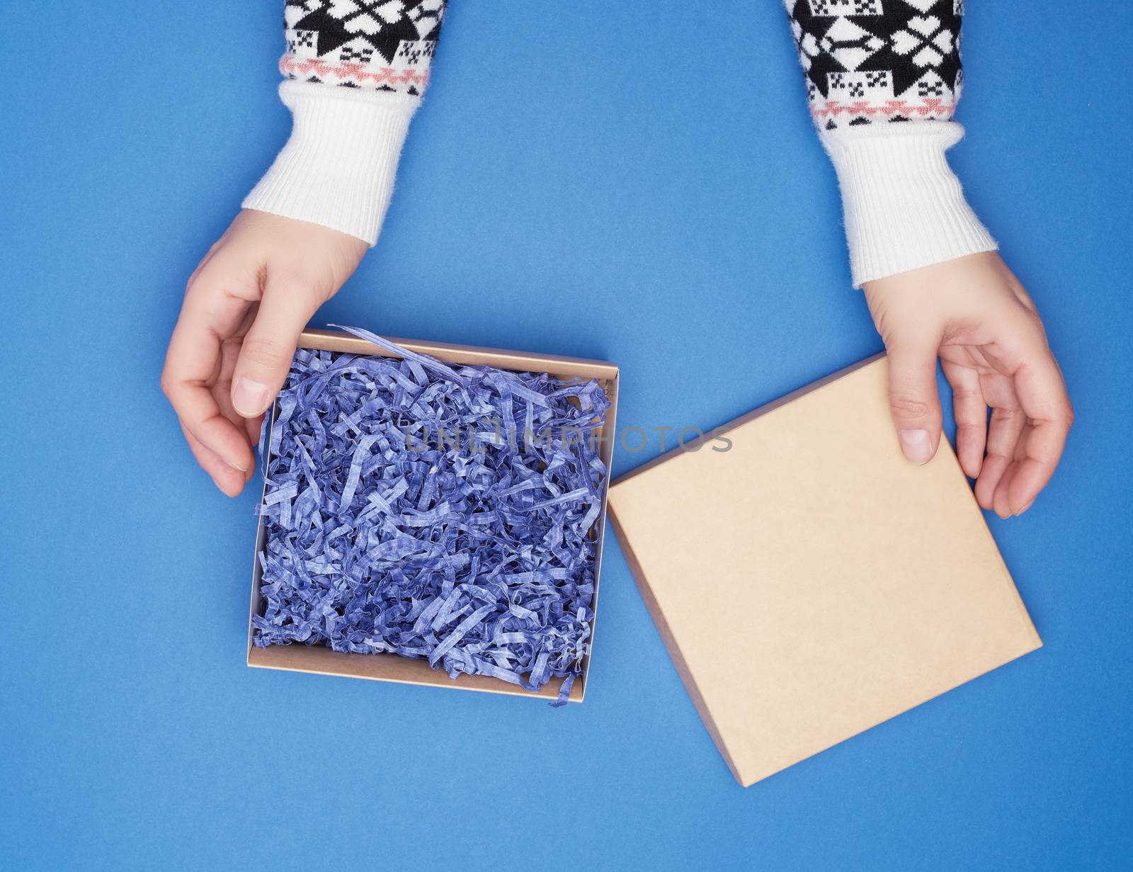 two female hands in a white sweater hold open square brown box on a blue classic background, top view