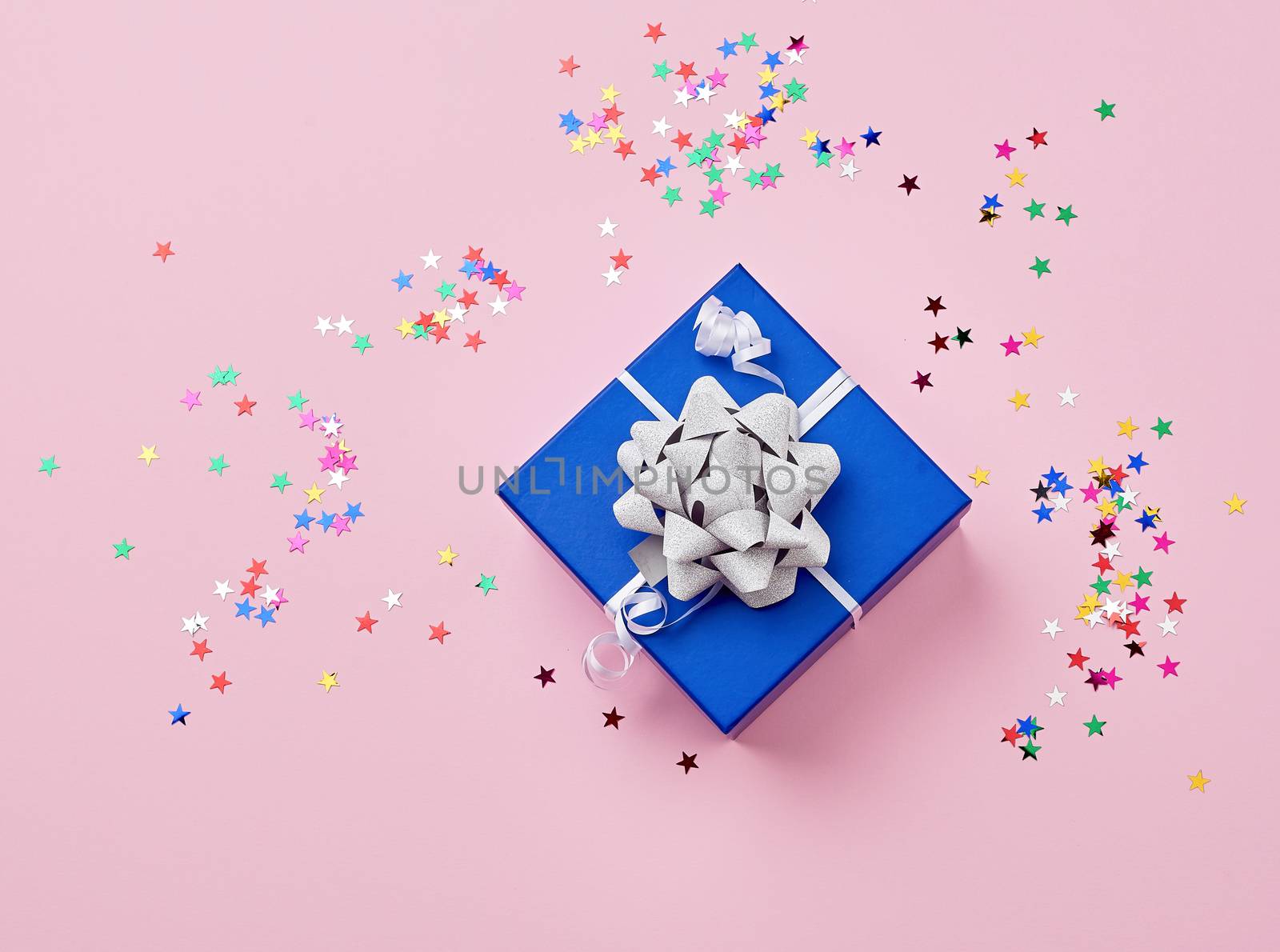 blue cardboard square box tied up with a white ribbon with a shiny bow and scattered multicolored shiny star-shaped confetti on a pink background, holiday backdrop, top view