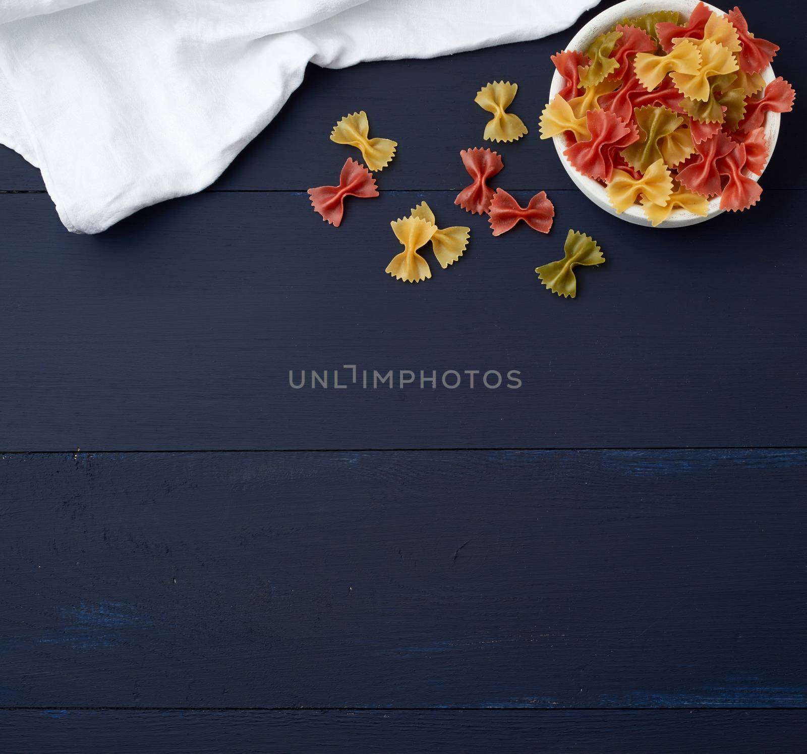 raw pasta in the form of bows in a wooden round plate and white textile napkin on a blue wooden background, top view. Vegetarian nutrition. Italian cuisine