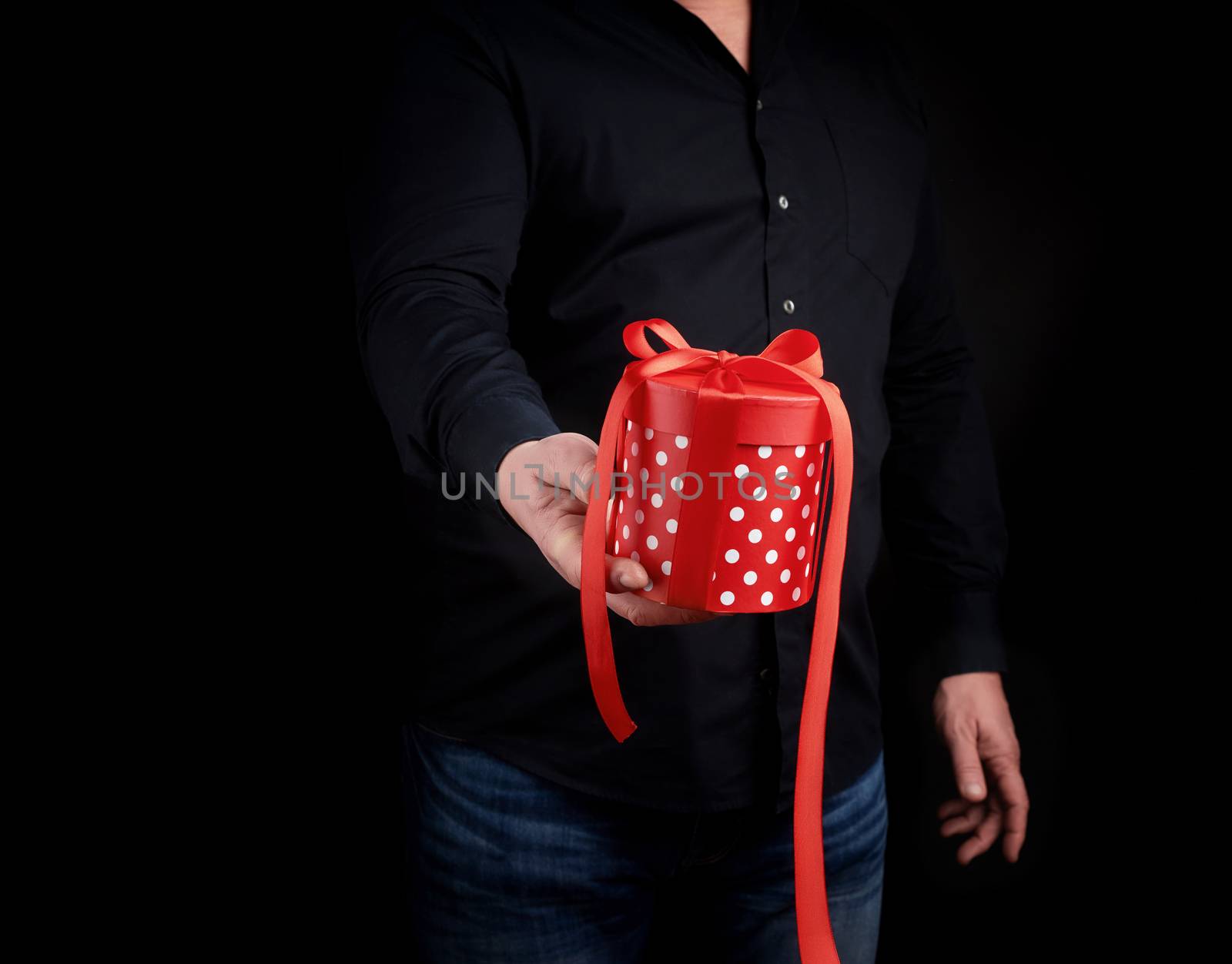 adult man in a black shirt holds in his hand a round red box of polka dots with a red bow on a black background, concept of a holiday greetings, surprise and gratitude