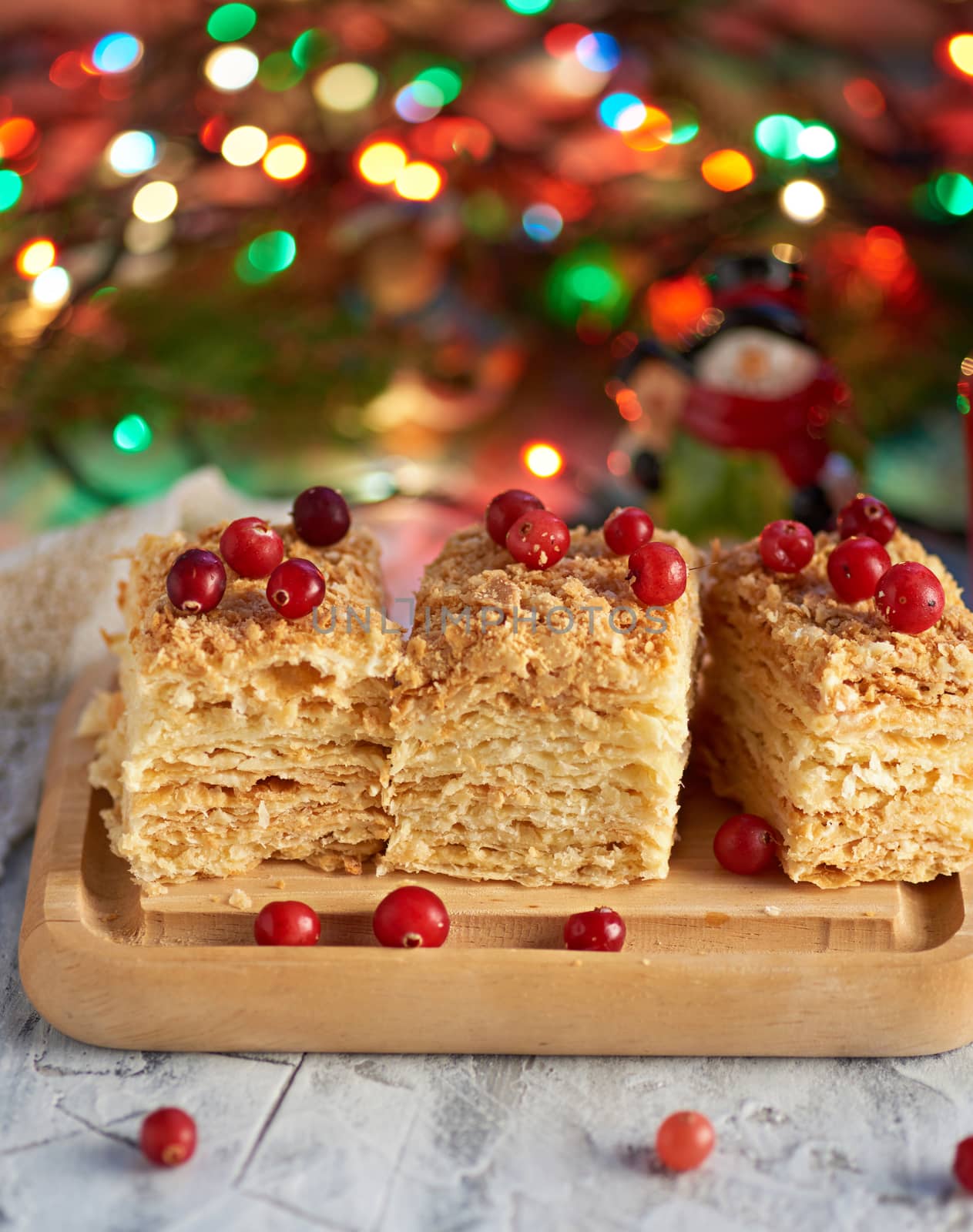 pieces of baked layer of cake Napoleon with butter cream on a wooden board, behind a burning Christmas garland, festive backdrop