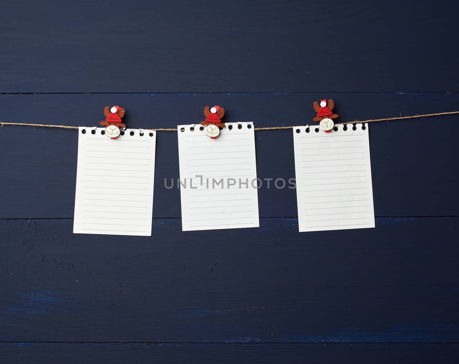 empty white notebook paper sheets hanging on decorative holiday clothespins, blue wooden background. Clean sheet, template. Design element.