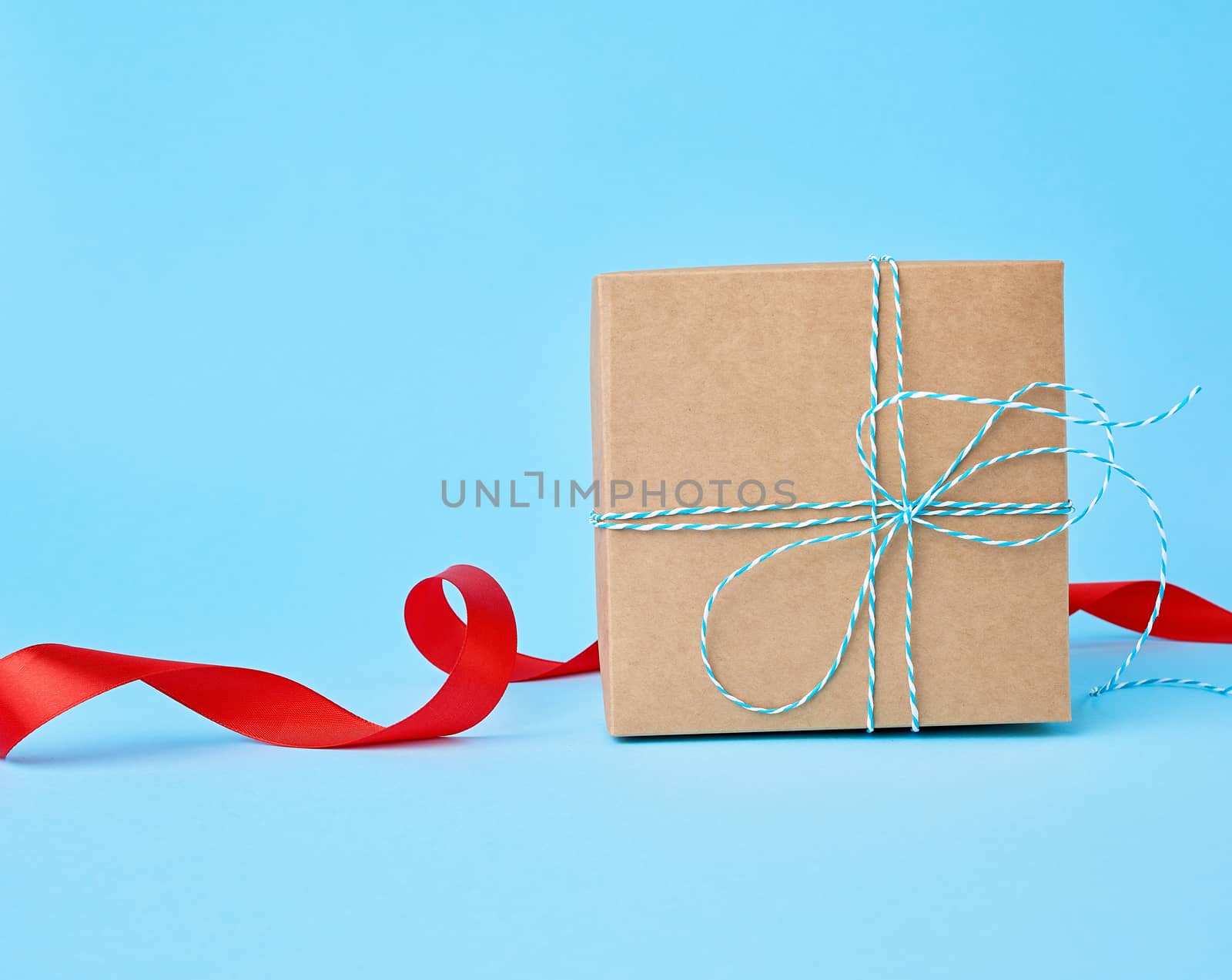 square brown cardboard box for a gift and twisted silk red ribbon on a blue background. Beautiful box gift, great design for any purposes. 