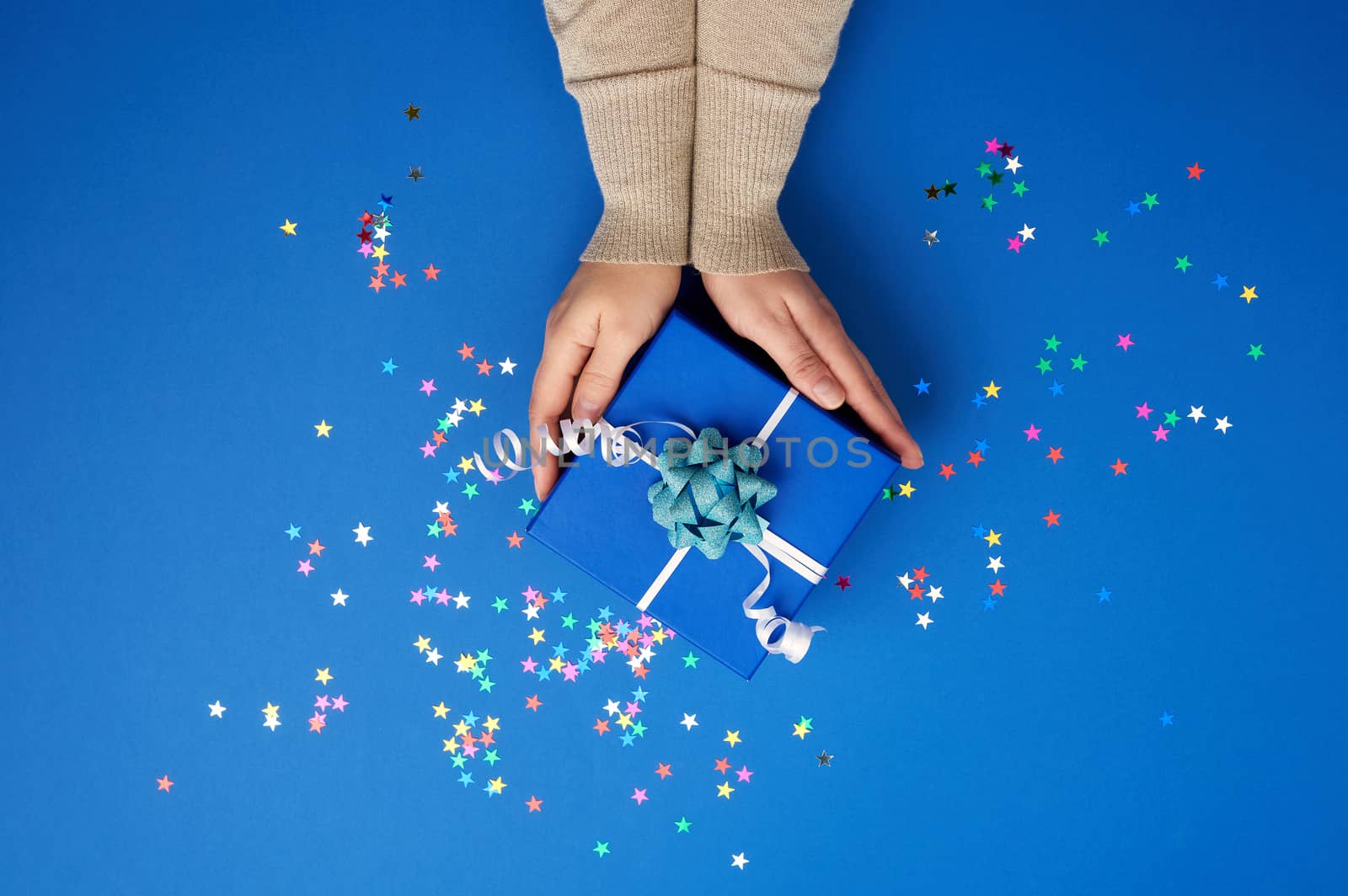 two female hands in a golden sweater hold closed square blue box with a bow on a blue classic background, top view, festive backdrop