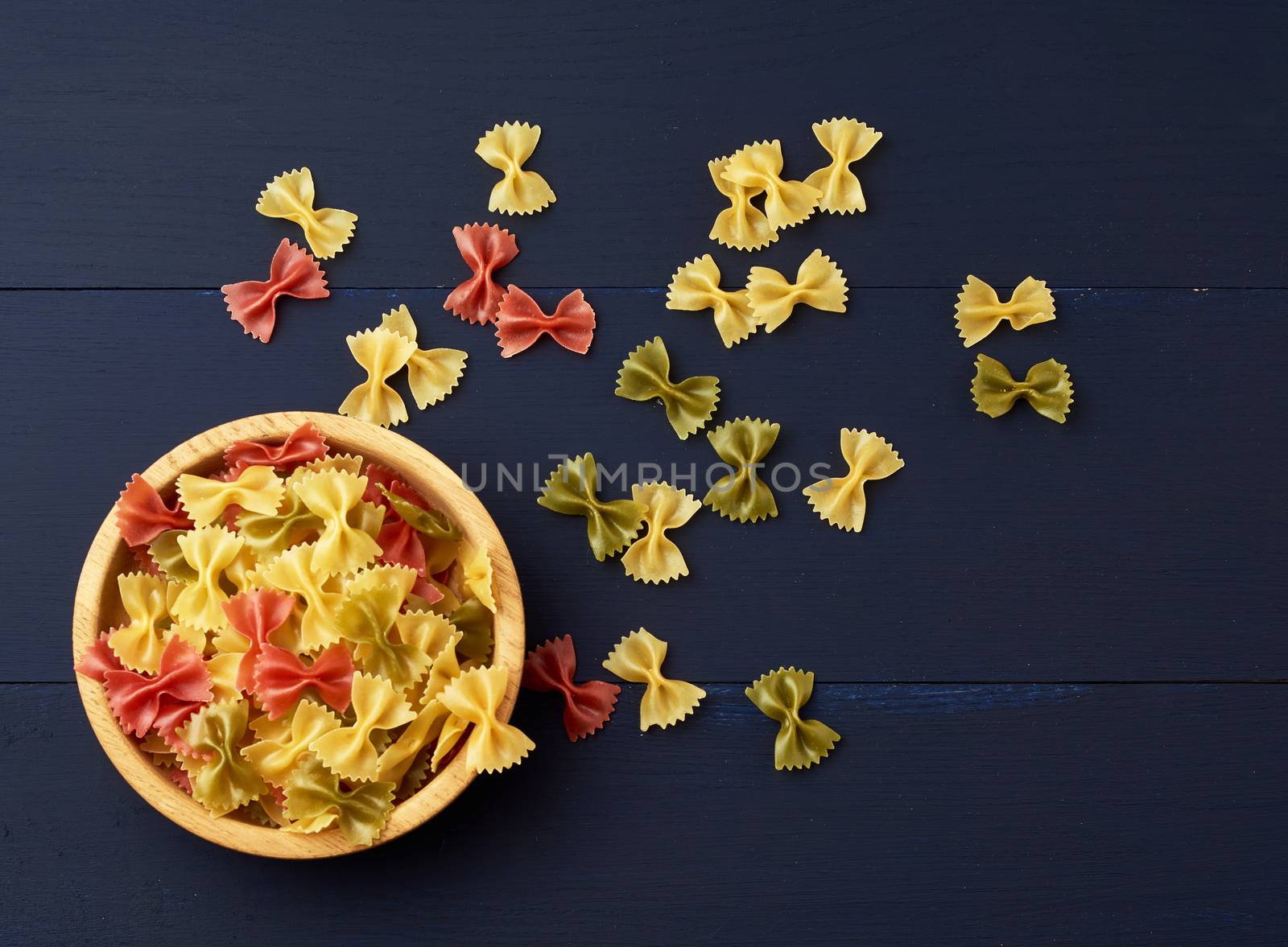 raw pasta in the form of bows in a wooden round plate and white textile napkin on a blue wooden background, top view. Vegetarian nutrition. Italian cuisine, copy space