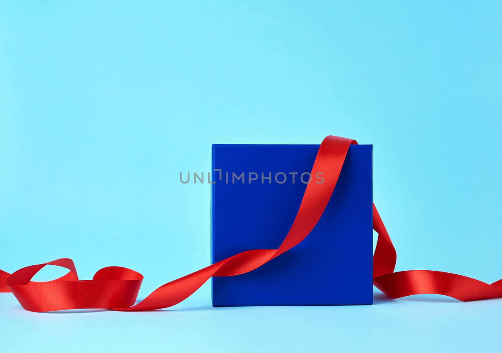 square blue cardboard box for a gift and twisted silk red ribbon on a blue background. Beautiful box gift, great design for any purposes. 