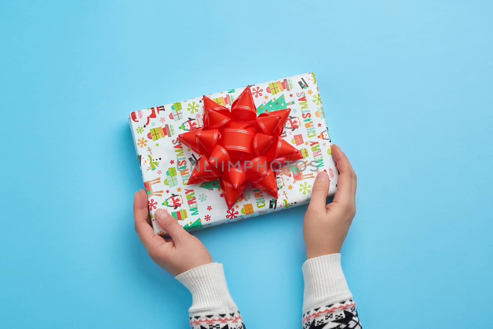 two female hands hold a paper closed box with red bow on a blue background. Festive concept