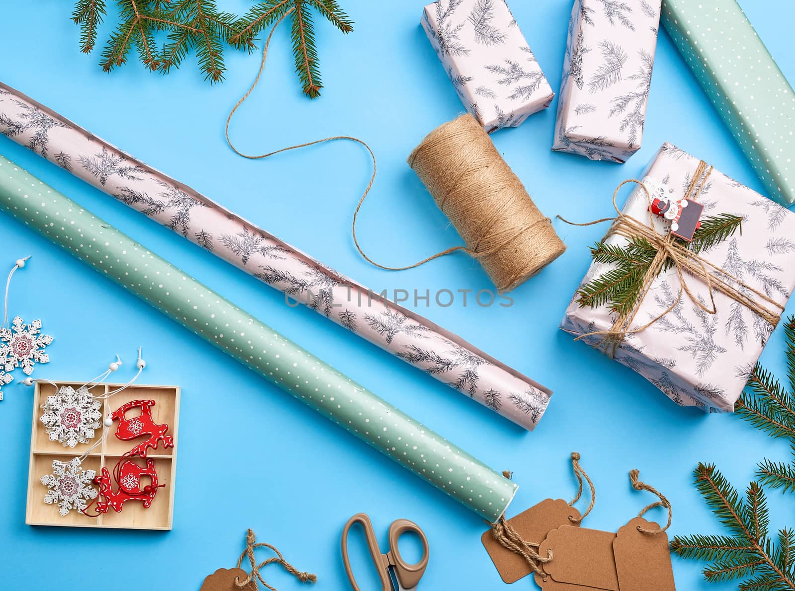 rolls with wrapping paper, brown rope, scissors, decor and a wrapped square box with a gift. DIY gift making process, blue background