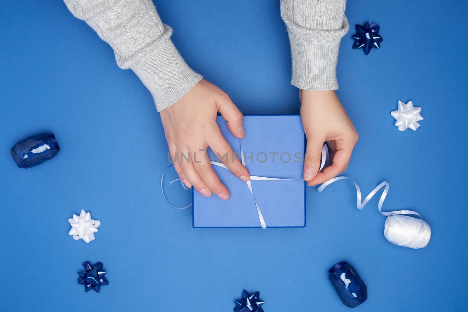 two female hands in a gray sweater hold closed square blue box with a bow on a blue classic background, top view, festive backdrop