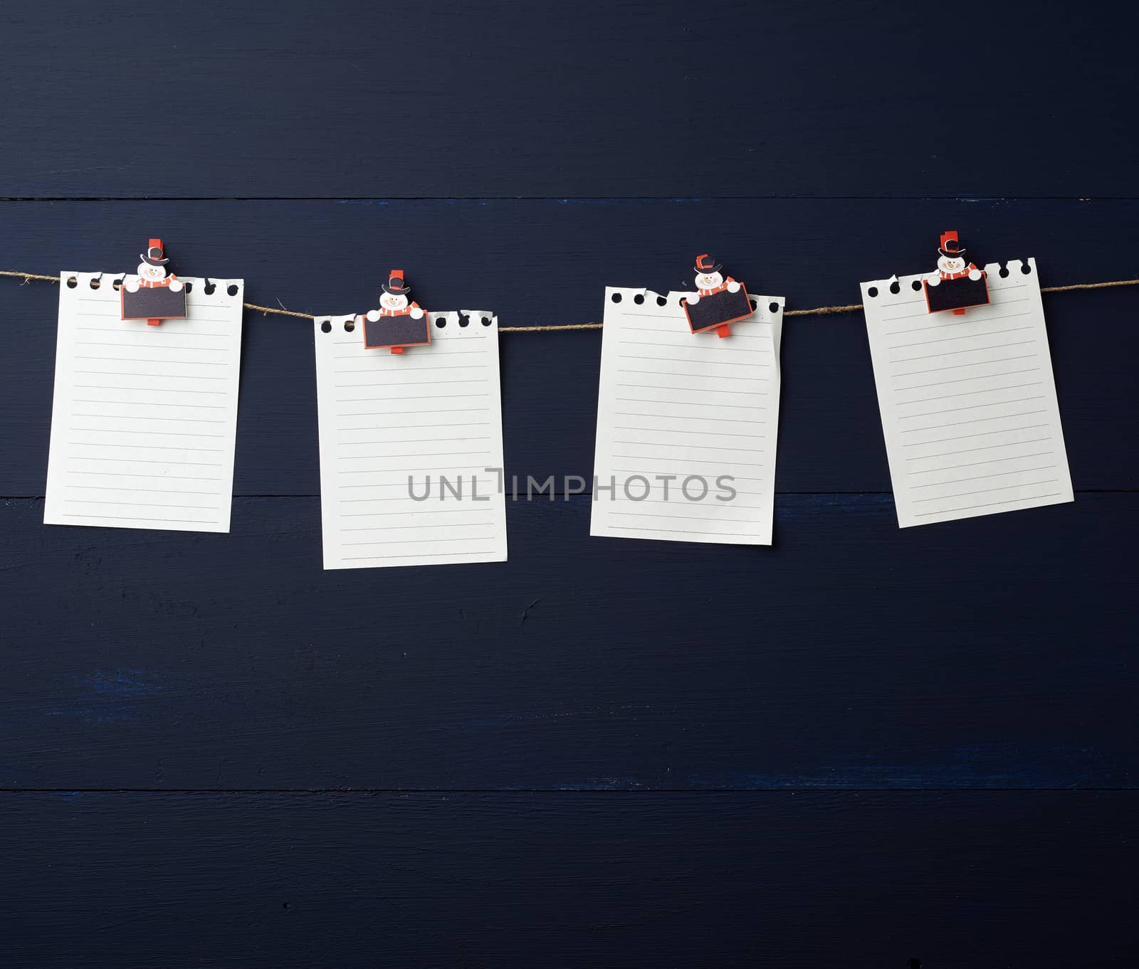 empty white notebook paper sheets hanging on decorative holiday clothespins, blue wooden background. Clean sheet, template. Design element.