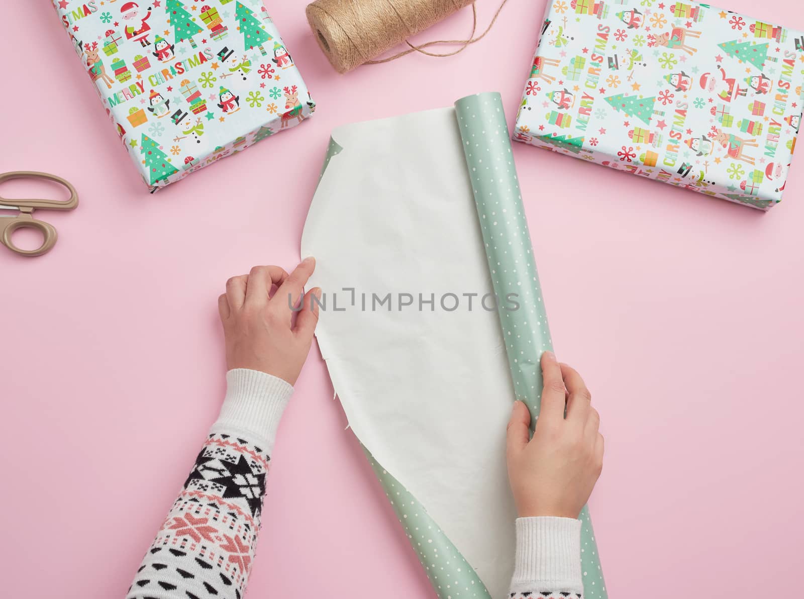 two female hands unfold a roll of wrapping paper, next to two gifts, pink background, preparing surprises