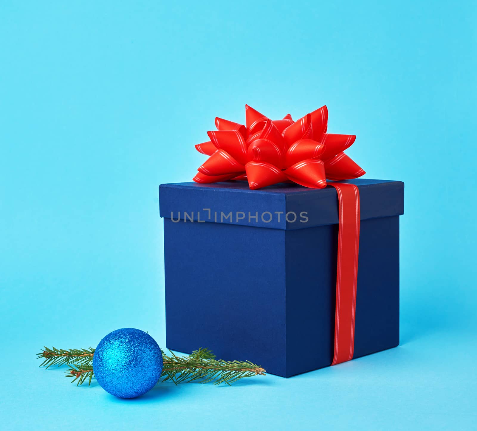 square blue cardboard box with a red bow, sprig of spruce with a shiny blue ball on a blue background, festive Christmas background. Christmas greeting card