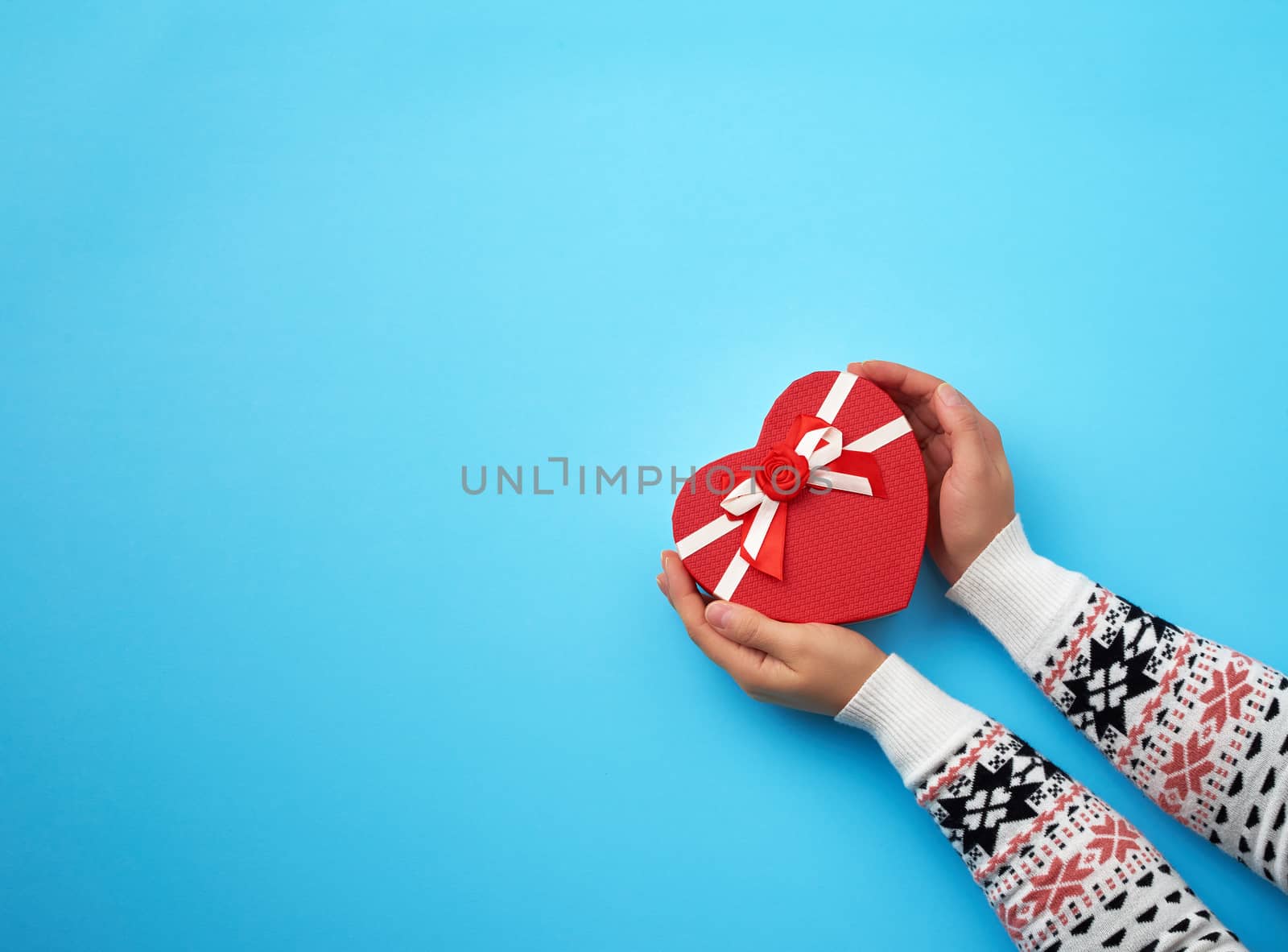 two female hands hold a paper closed red box in the shape of a heart on a dark blue background, festive backdrop, copy space