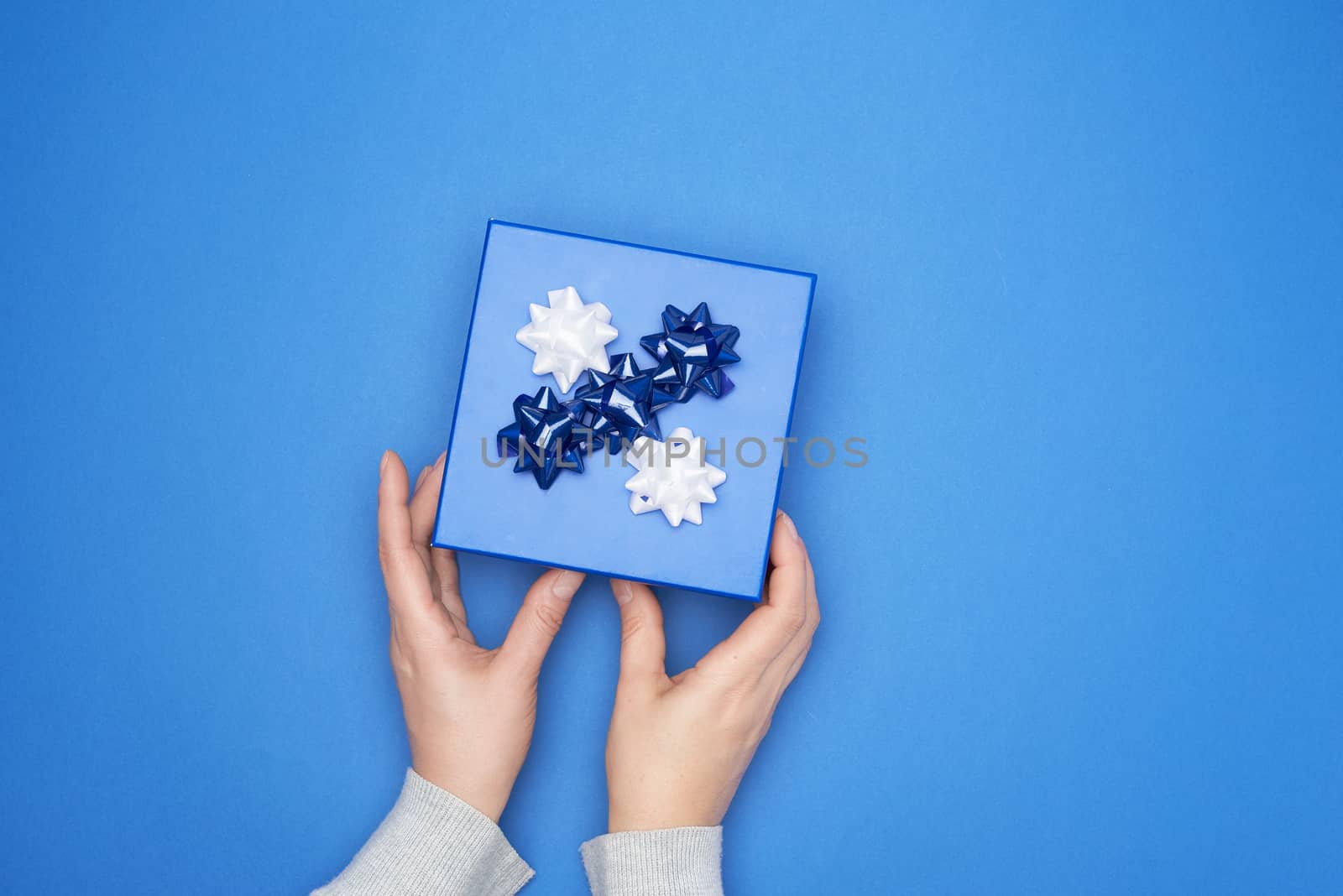 two female hands in a gray sweater hold closed square blue box with a bow on a blue classic background, top view, festive backdrop