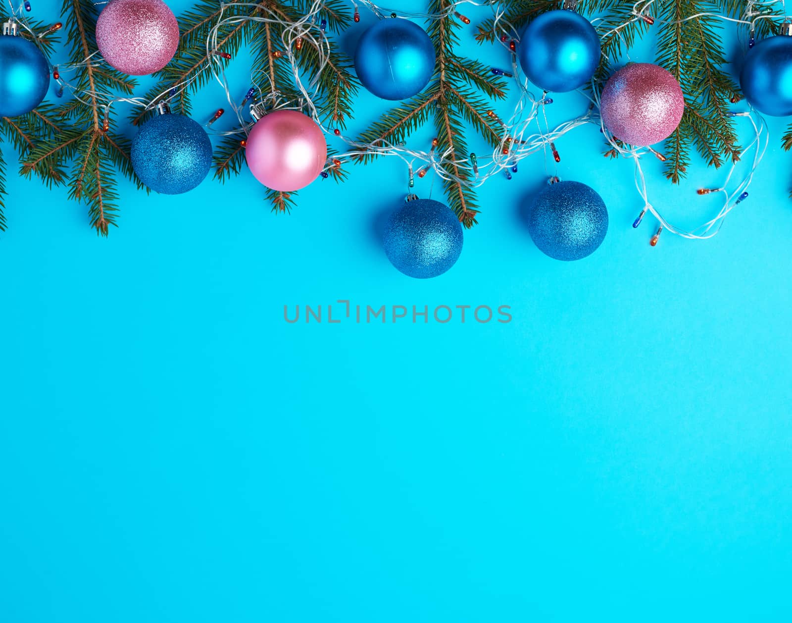 burning multi-colored light bulbs on a garland with a white cable and green twigs, blue and pink balls, blue background, copy space