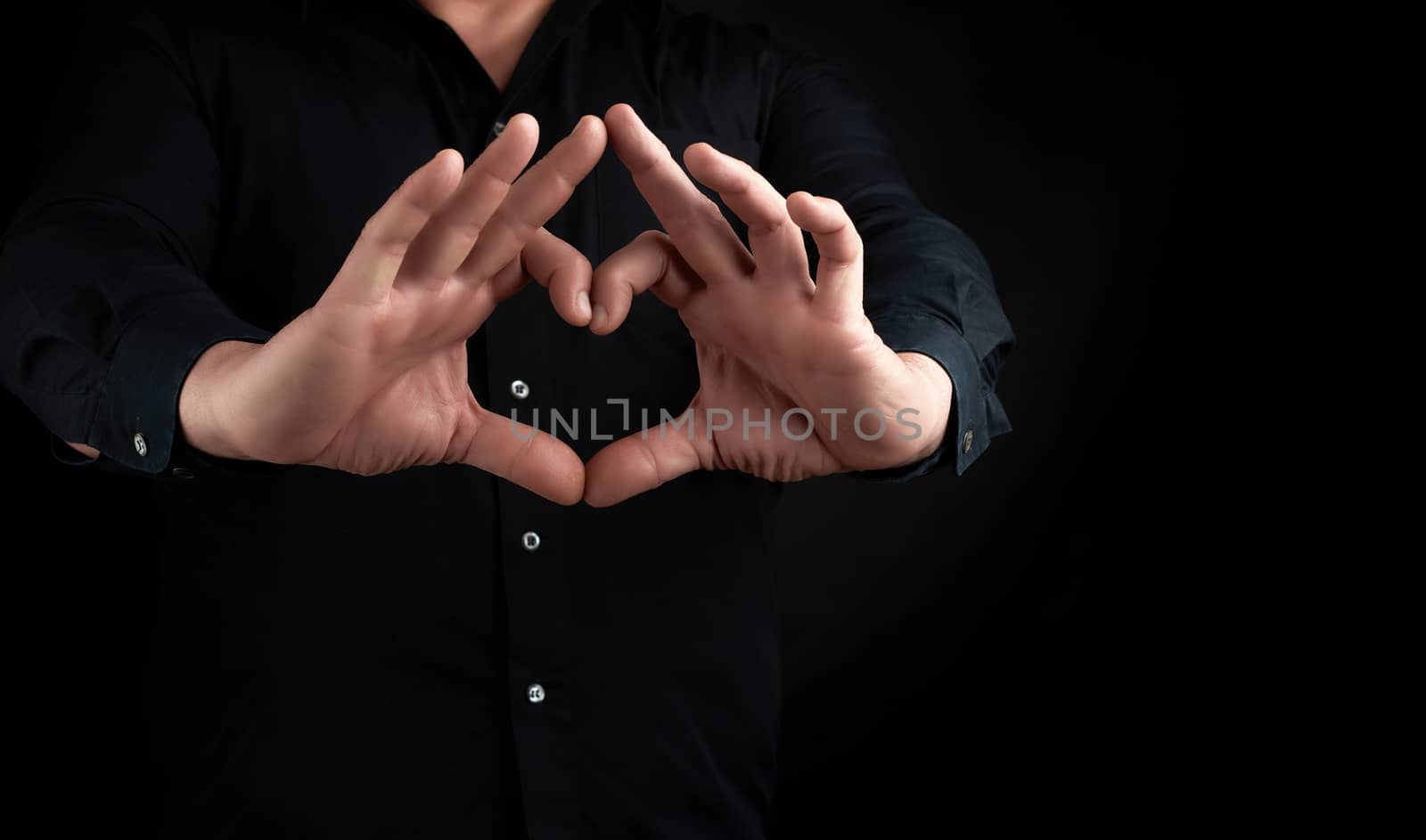 adult man in a black shirt shows hands a heart gesture on a dark background, concept of love and peace