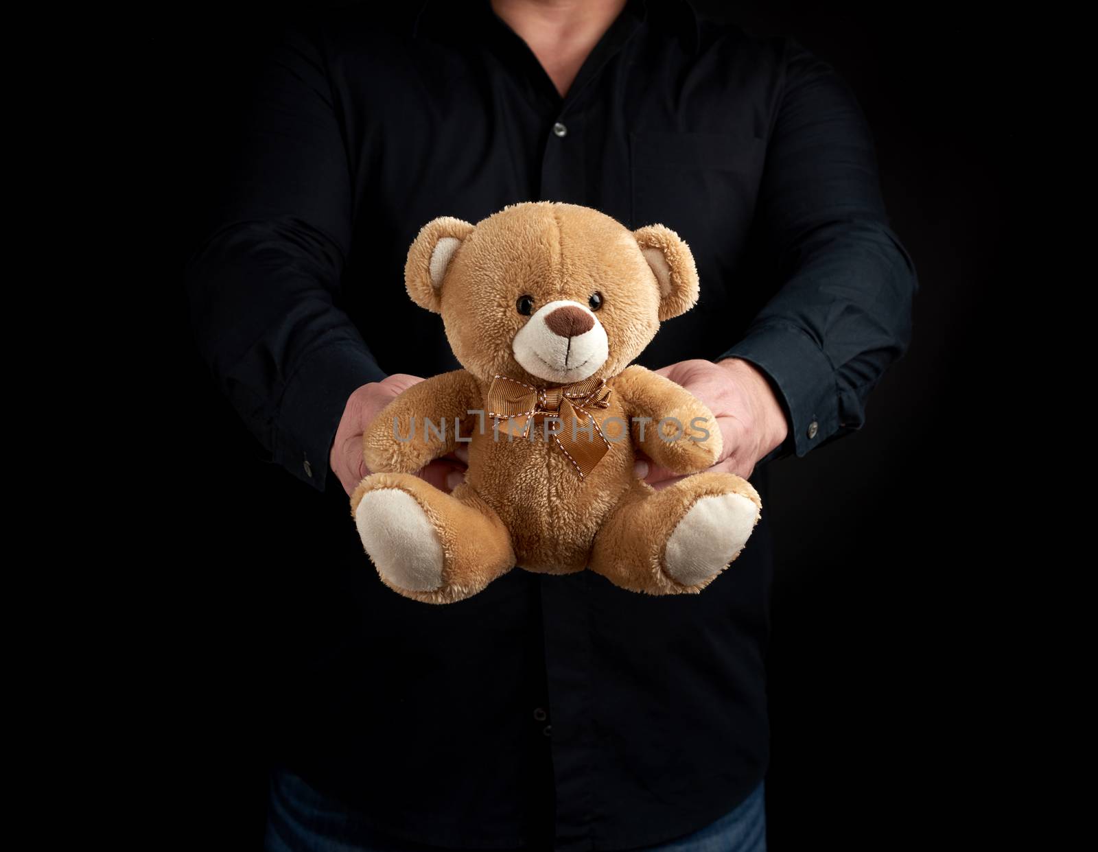 adult man in a black shirt holds a brown teddy bear on a dark background, concept of congratulations on the holiday of Valentine's Day on February 14, low key