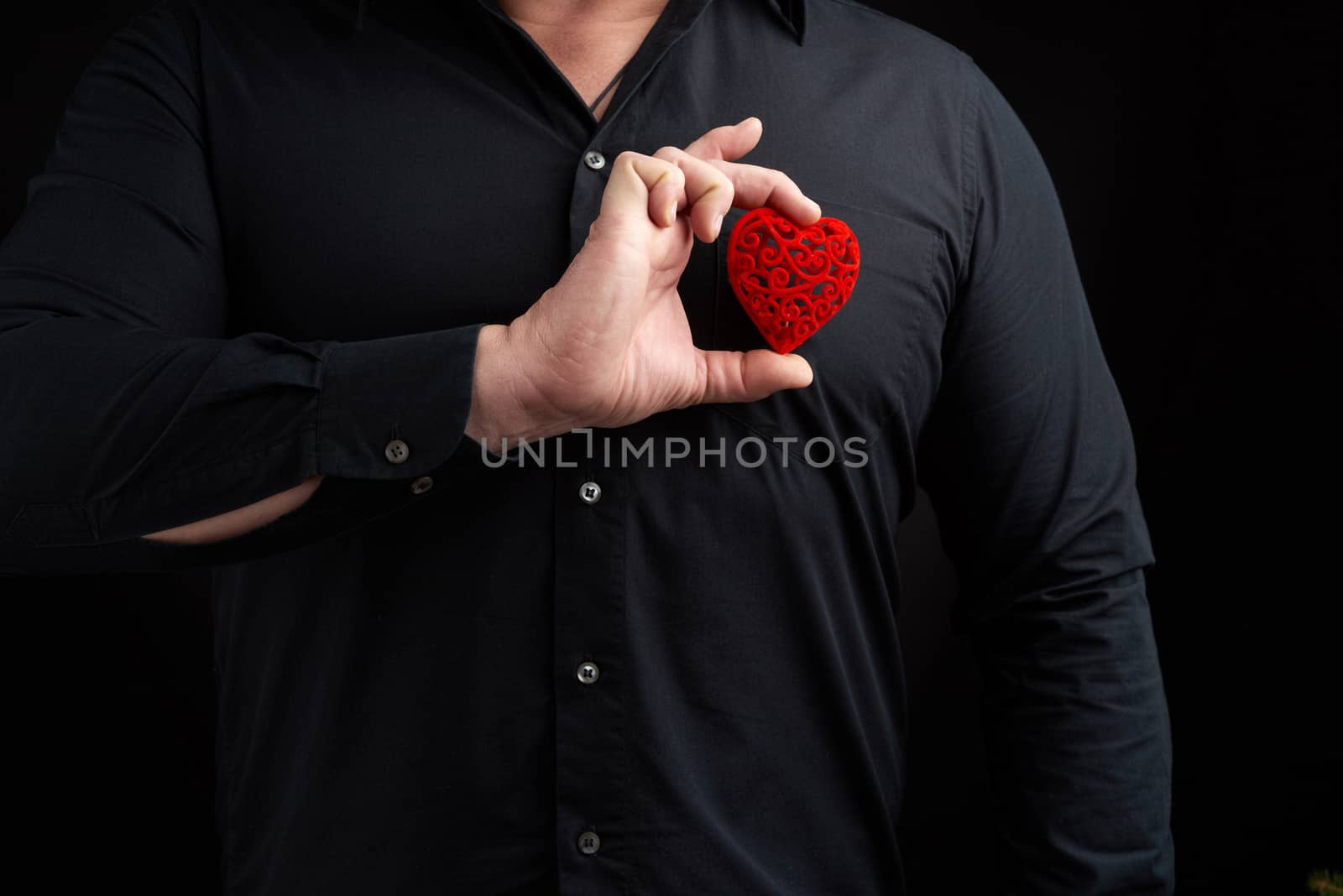 adult man stands on a dark background wearing a black shirt and holding a red carved heart near his chest in his right hand, concept of love