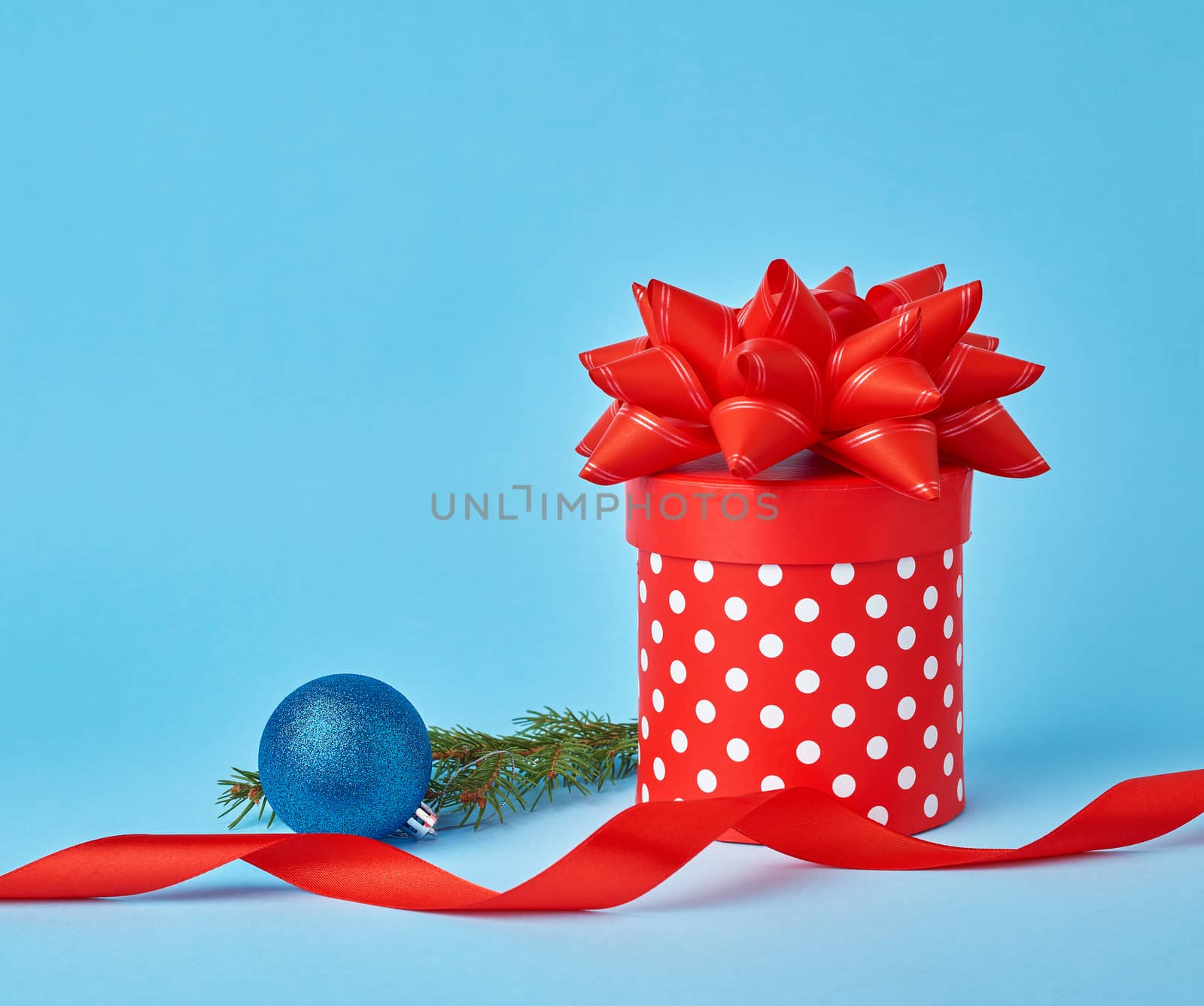 round red cardboard box in white polka dots with a bow, sprig of spruce with a shiny blue ball on a blue background, festive Christmas background