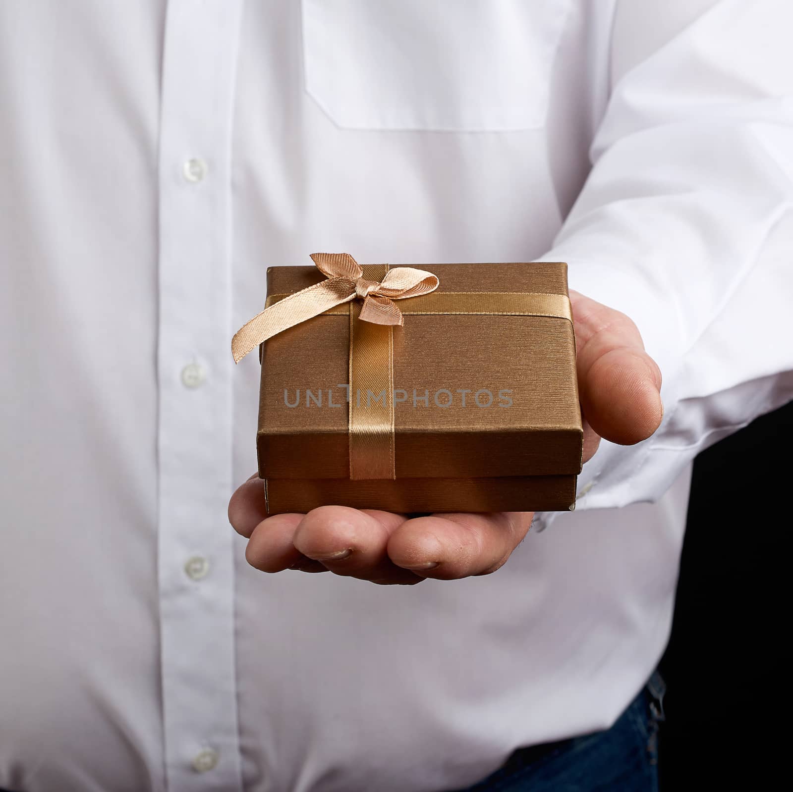 adult man in a white shirt holds in his hand a square small box with a bow, concept of a proposal to get married, a dark background, close-up