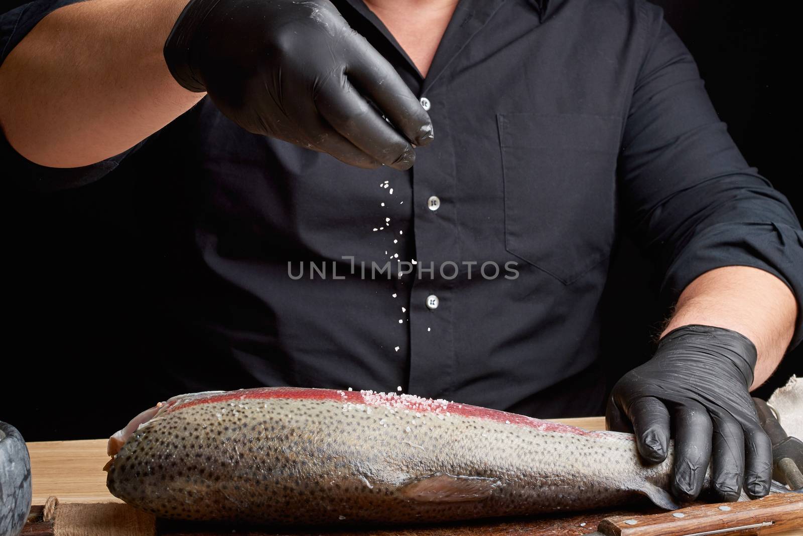 chef in a black shirt and black latex gloves prepares salmon fillet on a wooden cutting board, process of sprinkling with spices and salt, low key