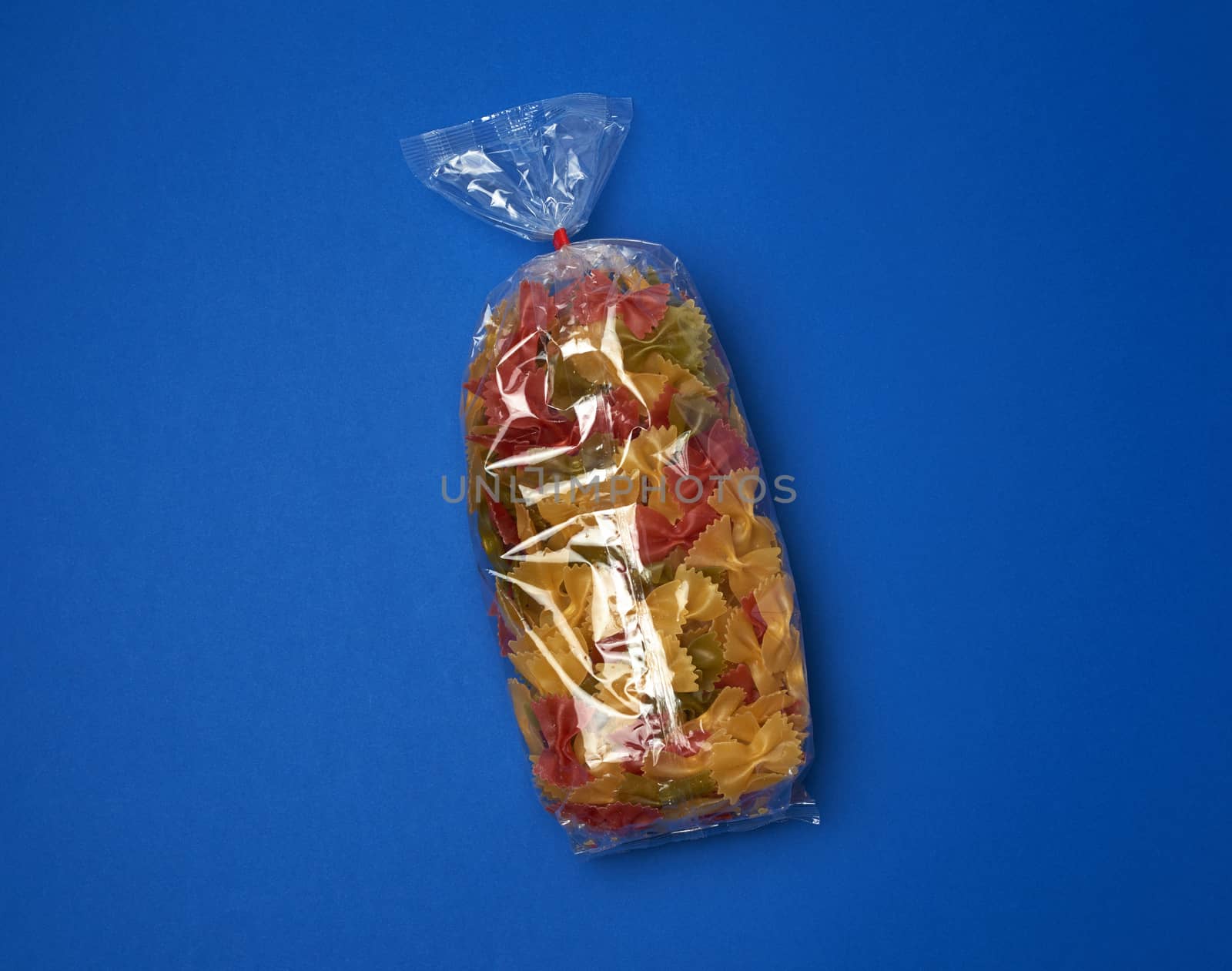 transparent cellophane bag with raw colorful pasta in the form of bows on a blue surface, copy space