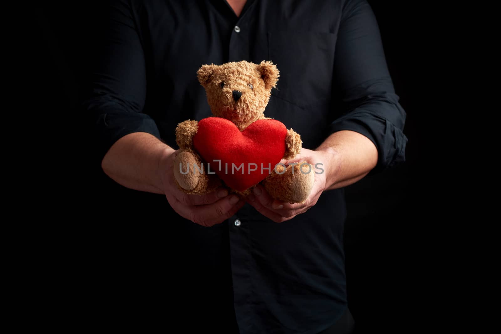 adult man in a black shirt holds a brown teddy bear with a red heart on a dark background,  concept of congratulations on the holiday of Valentine's Day on February 14, low key