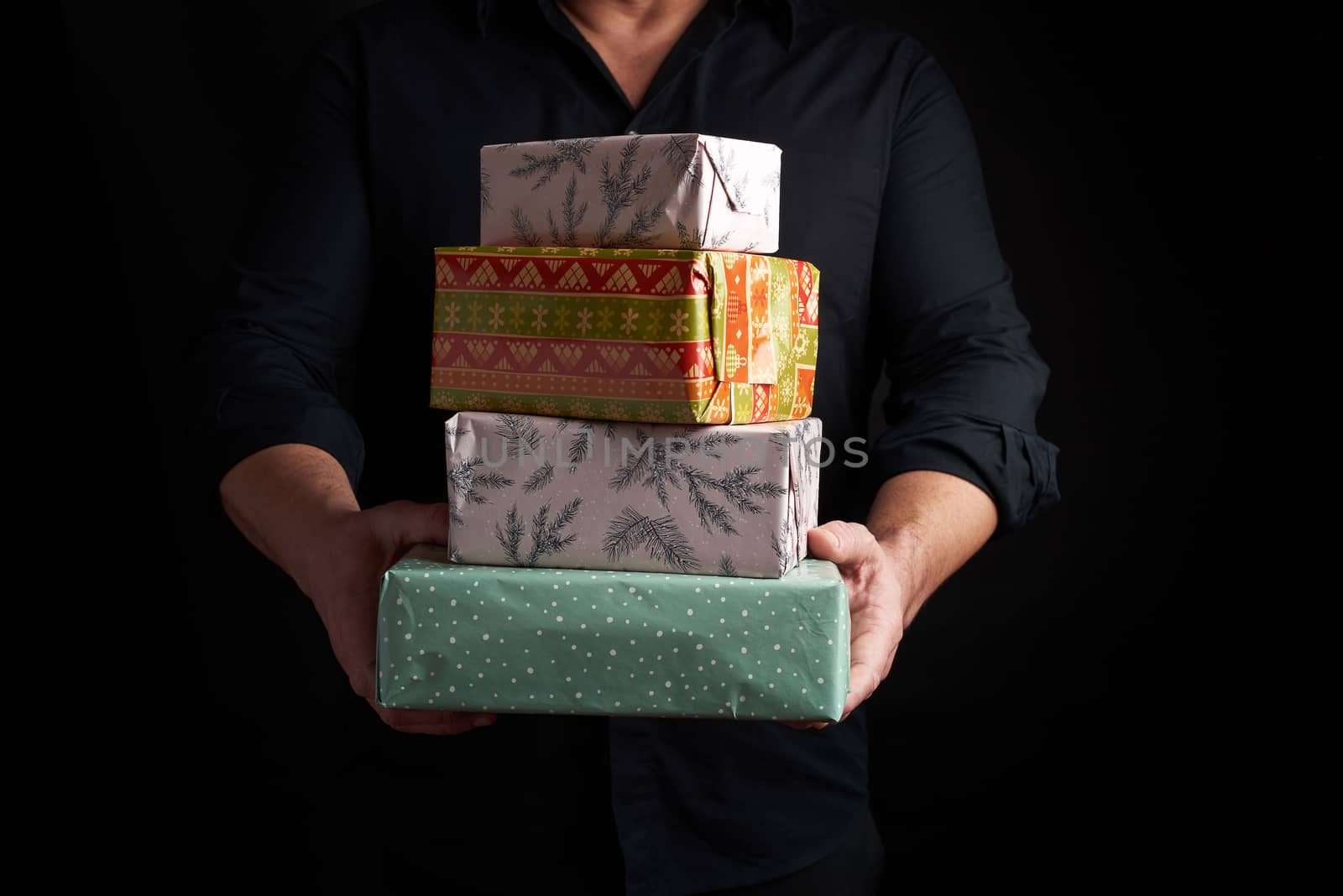 adult man in a black shirt holds in his hands a stack of paper-wrapped gifts on a black background, concept of holiday greetings, surprise and gift