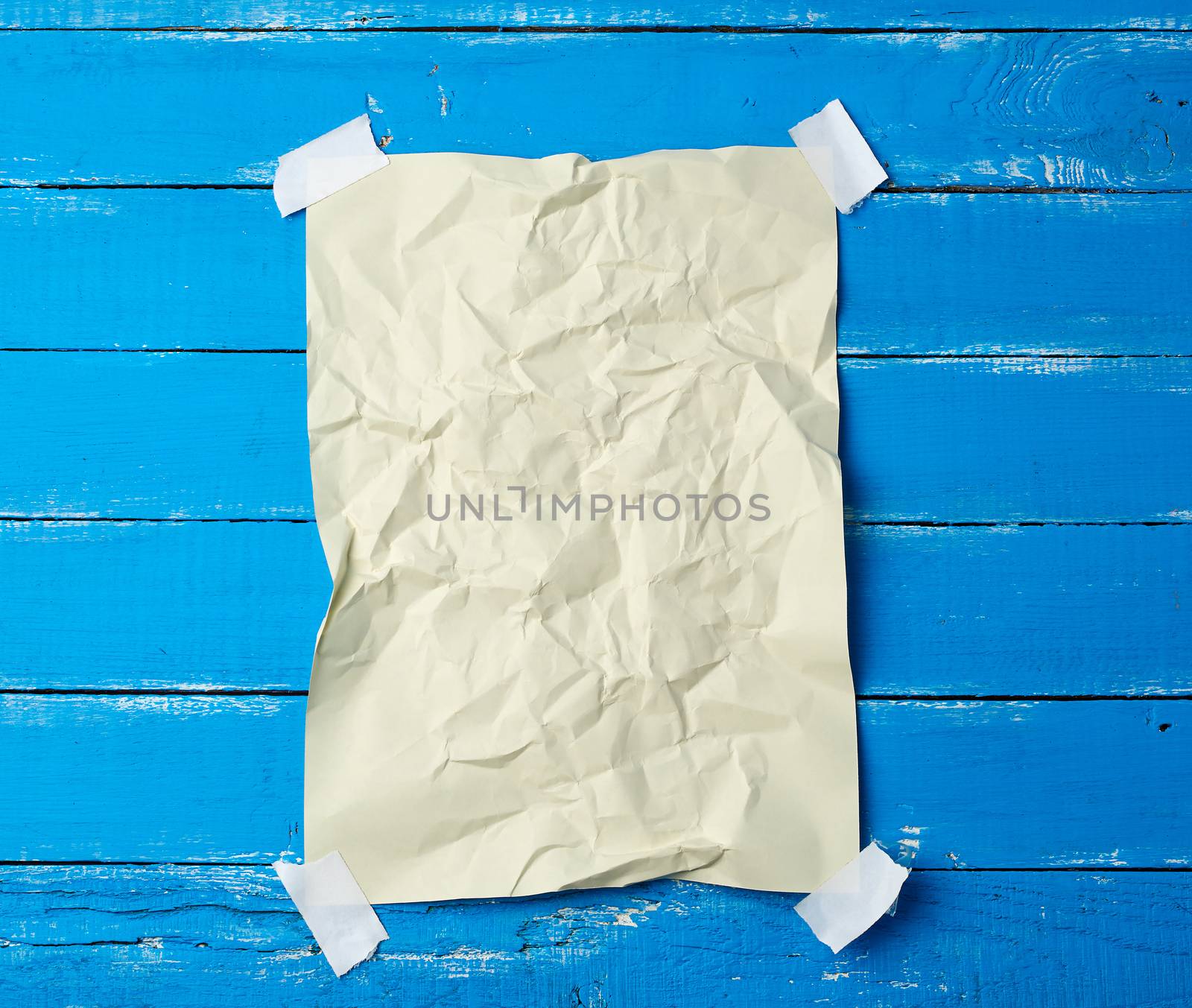 glued with adhesive tape in the corners of a rectangular empty beige crumpled sheet of paper on a blue wooden background, place for inscription