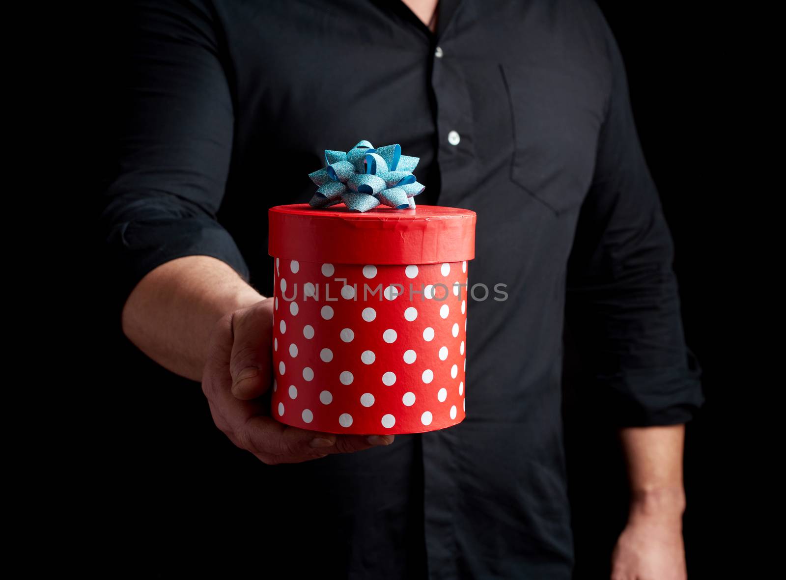 adult man in a black shirt holds in his hand a round red box of polka dots with a blue bow on a black background, concept of a holiday greetings, surprise and gratitude