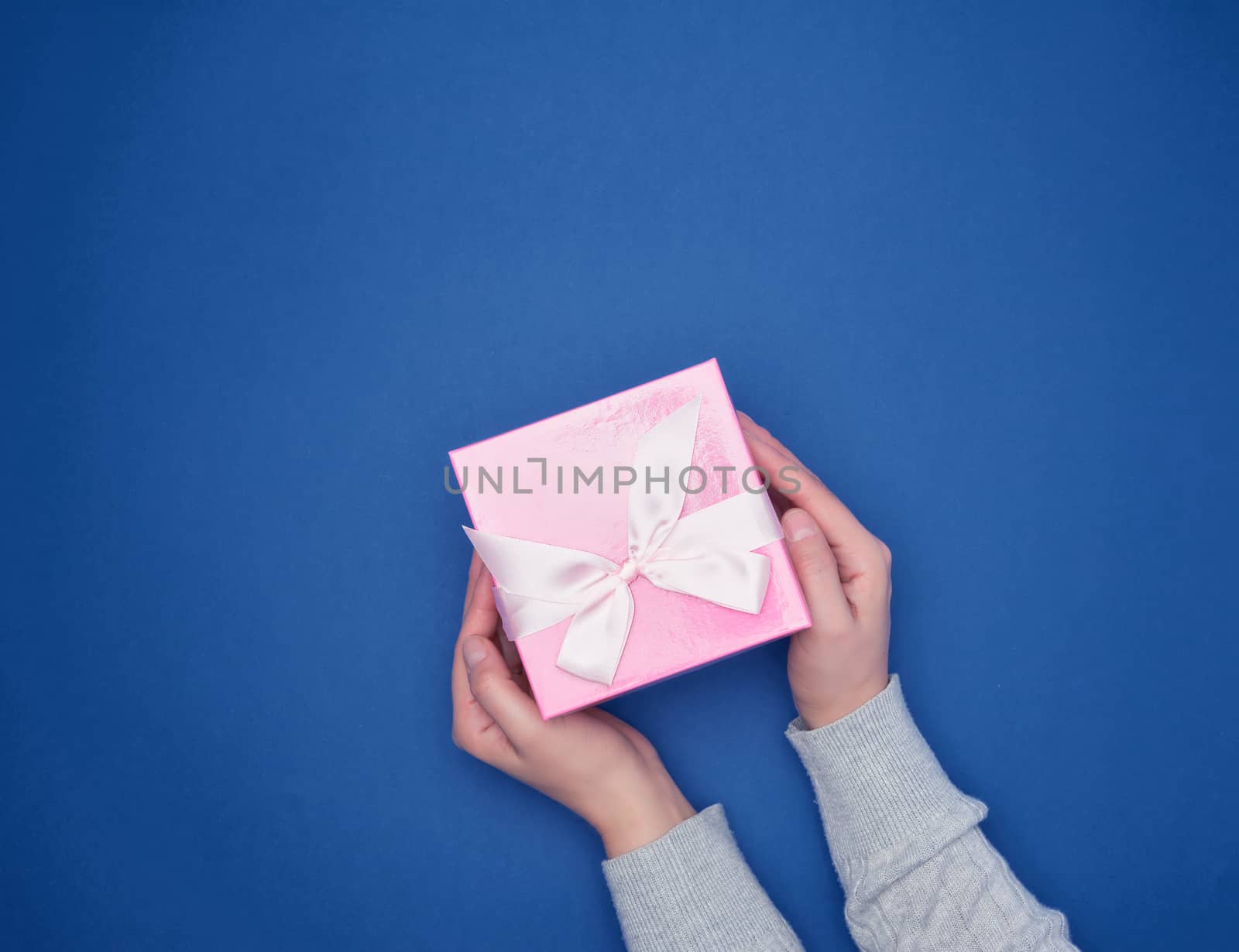 two female hands in a gray sweater hold a square pink box with a bow on a blue classic background, top view, festive backdrop