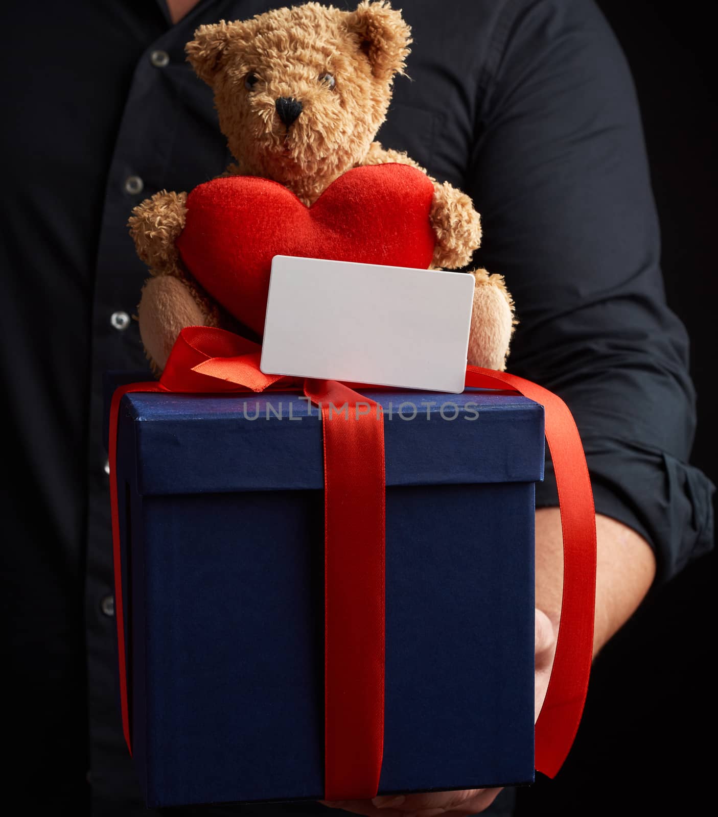 man in a black shirt holds a blue square box tied with a red ribbon and sits on top of a brown teddy bear with a heart, concept of congratulations on Valentine's Day on February 14