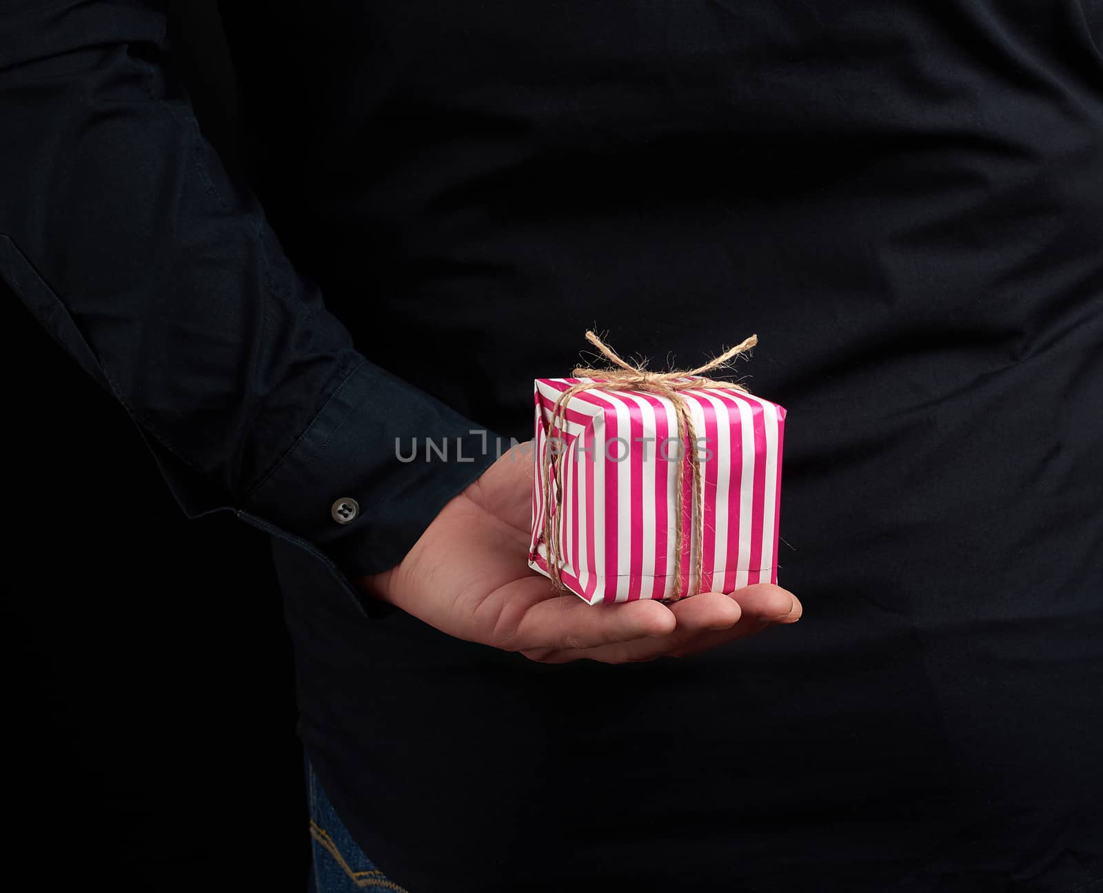 male hand holds a paper-wrapped box with a gift behind his back, man is wearing a black shirt, a concept of surprise