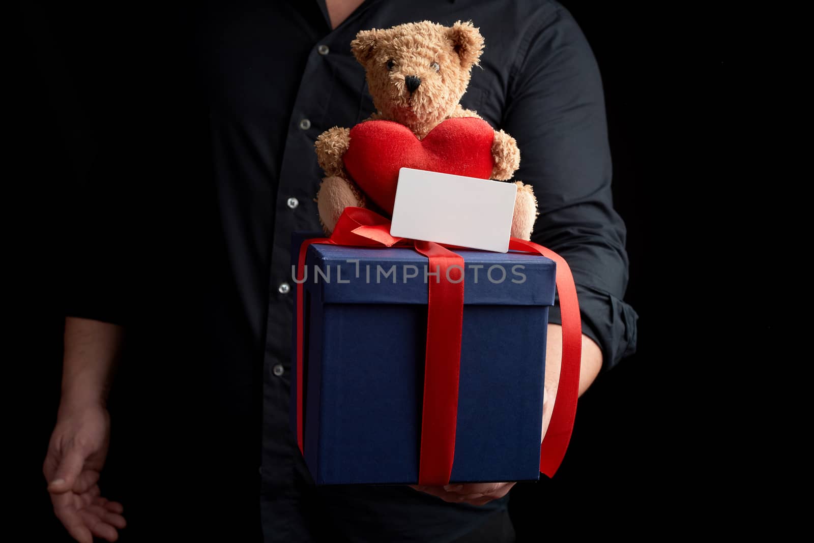 adult man in a black shirt holds a blue square box tied with a red ribbon and sits on top of a brown teddy bear with a heart, concept of congratulations on Valentine's Day on February 14