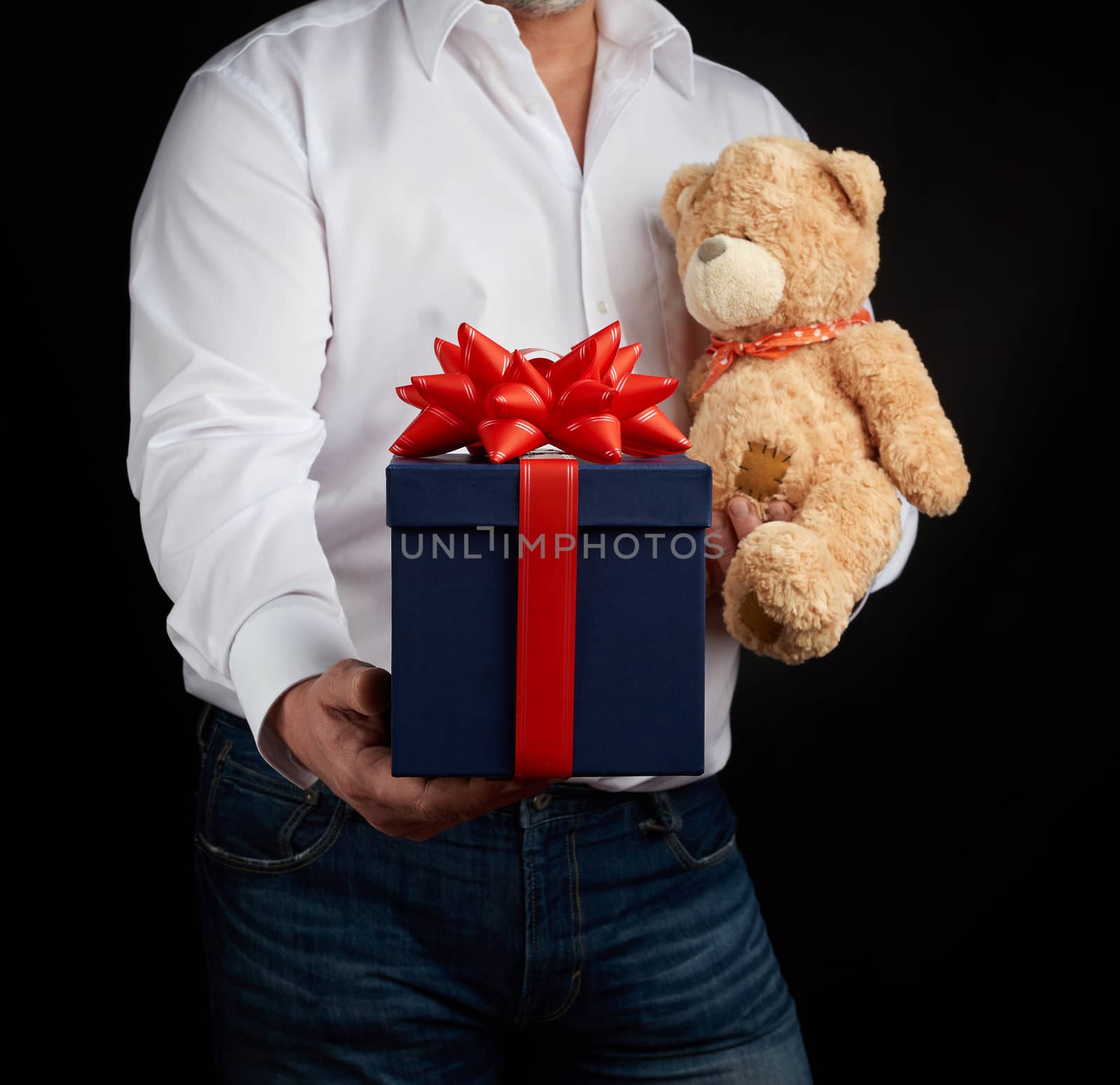 adult man in a white shirt holds a blue square box tied with a red ribbon and brown teddy bear, concept of congratulations on Valentine's Day on February 14