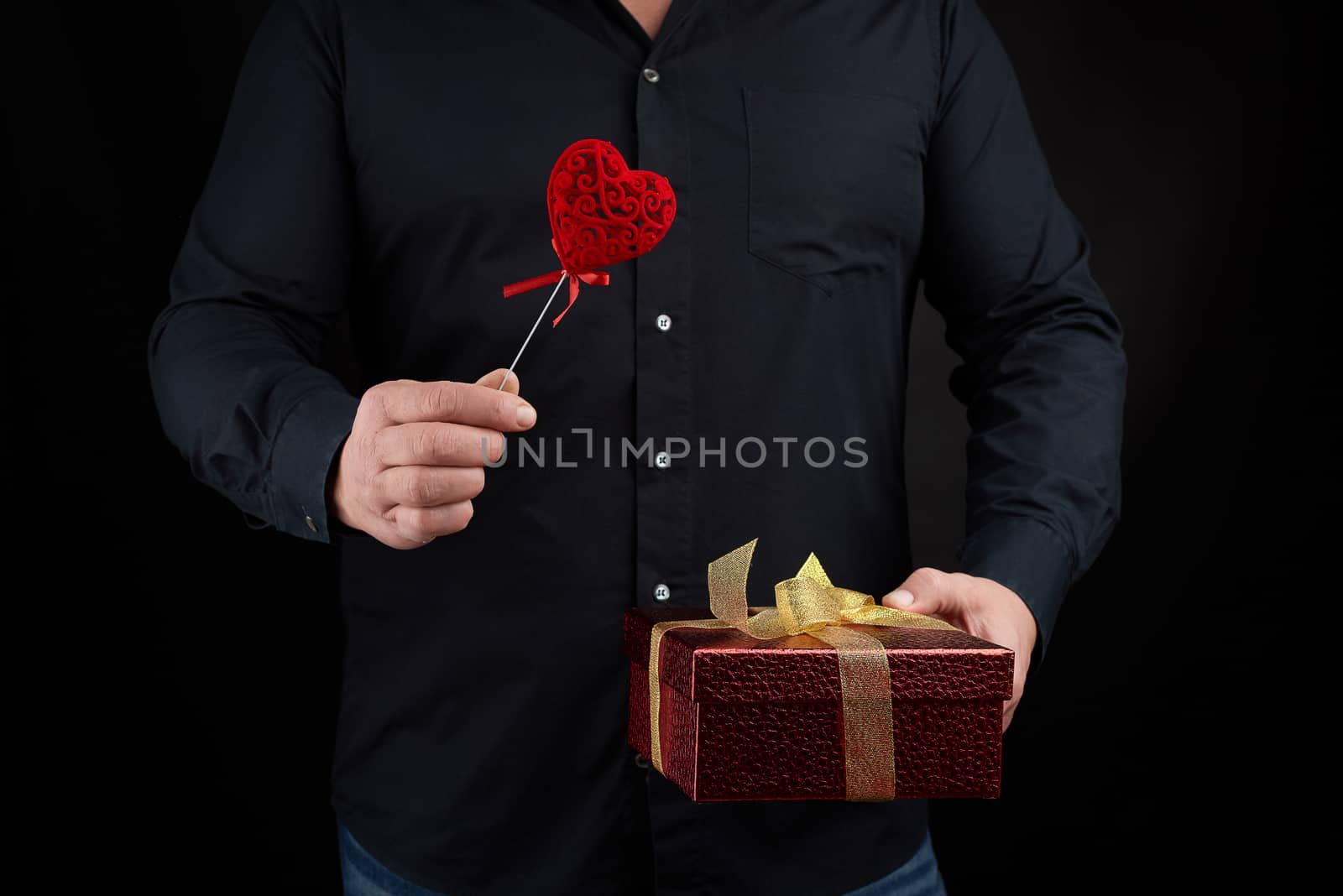 an adult man in a black shirt holds in his hand a red cardboard box tied with a golden bow and a red heart, holiday greetings concept, surprise