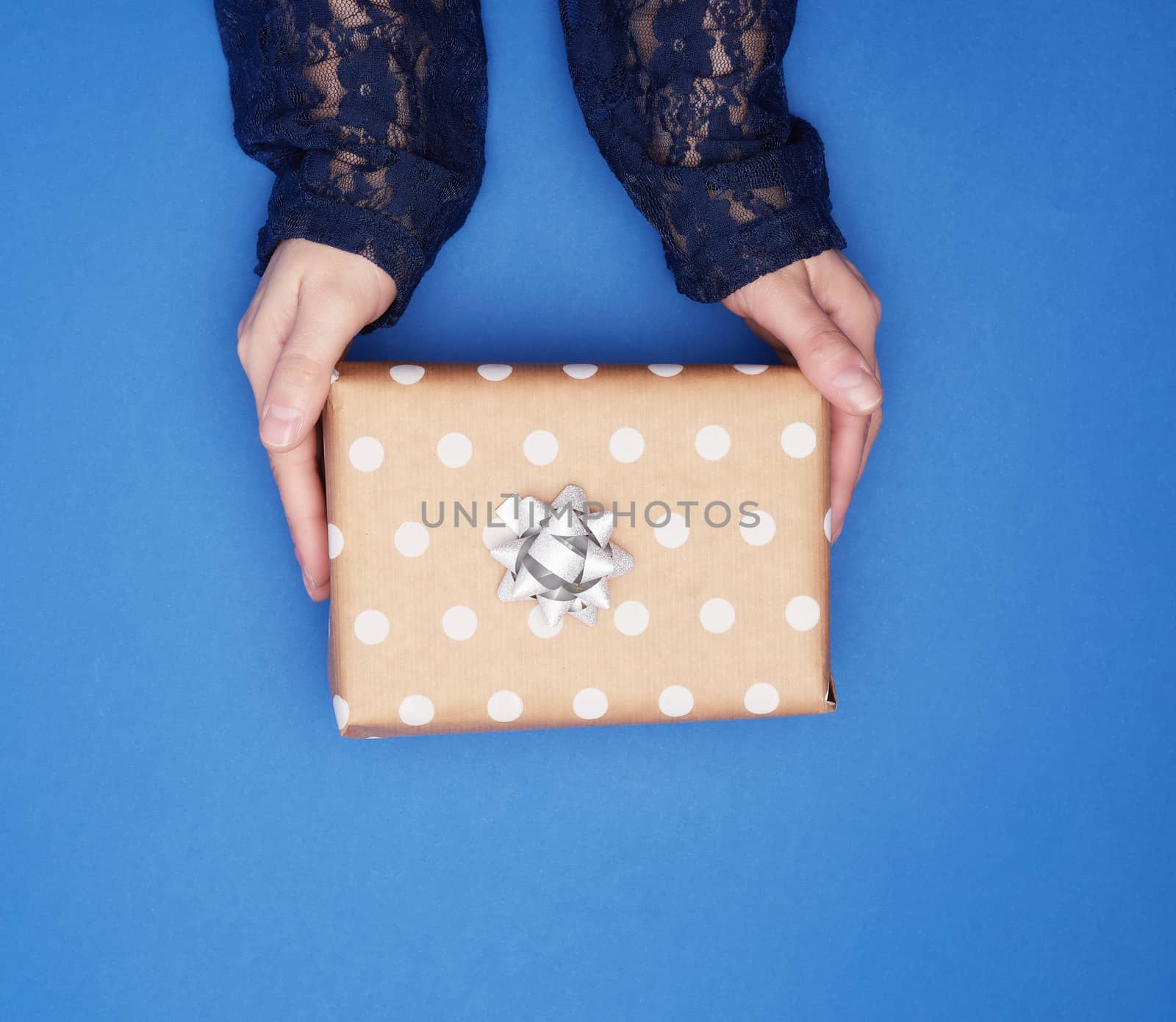 two female hands hold a square brown box with a bow on a blue classic background, top view, festive backdrop