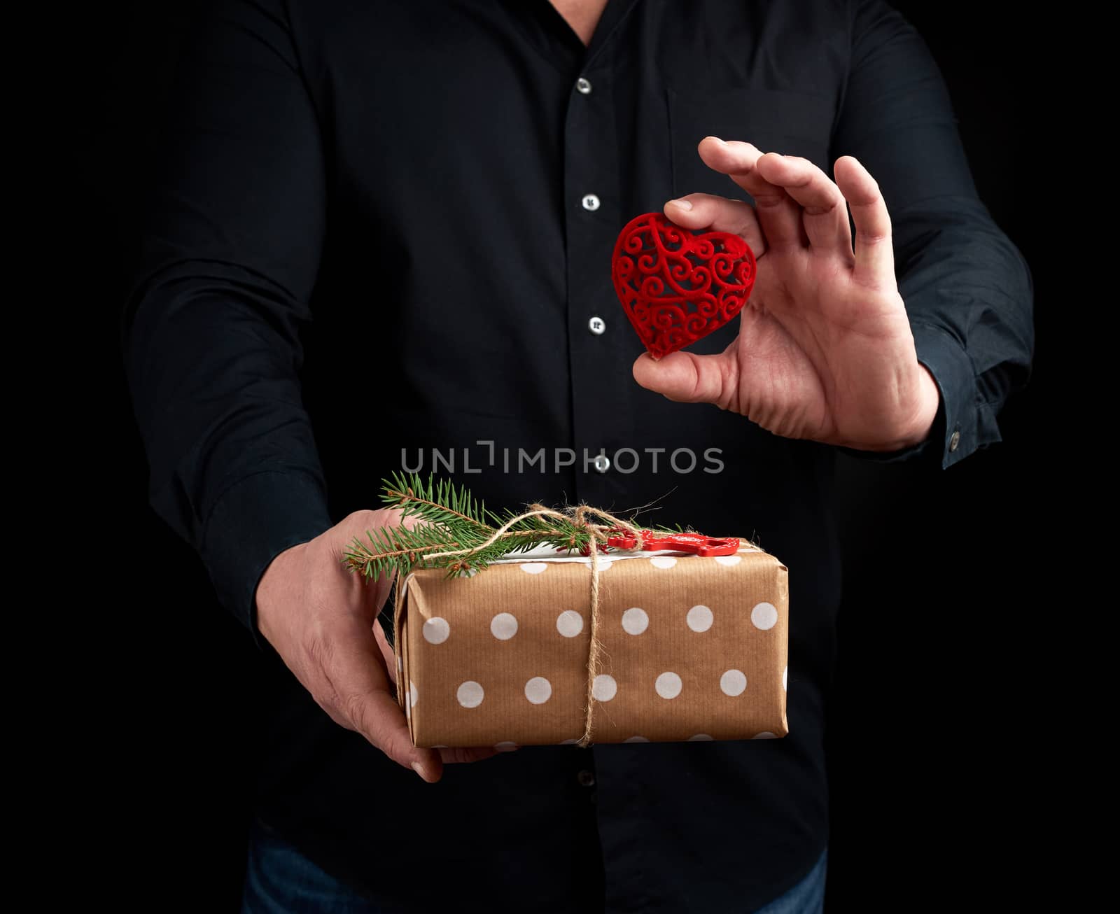 adult man is standing on a dark background wearing a black shirt and holding a wrapped gift in brown paper in his hand, and a red carved decorative heart in his left hand, concept of a holiday greetings