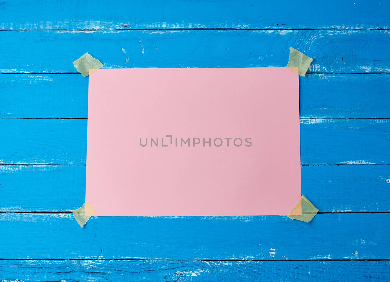 glued with adhesive tape a rectangular empty pink sheet of paper on a blue wooden background, place for an inscription