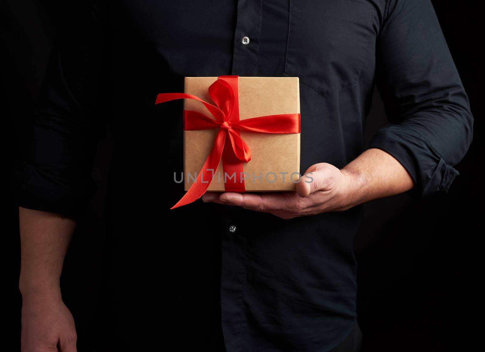 adult man in a black shirt holds in his hand a square brown boxlet tied with a red ribbon on a black background, concept of holiday greetings, surprise and gratitude