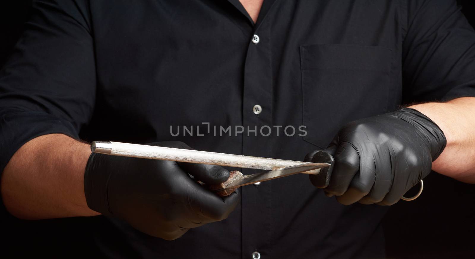 chef in a black shirt and black latex gloves sharpen a kitchen knife on an iron sharpener with a handle above the table, low key, close up
