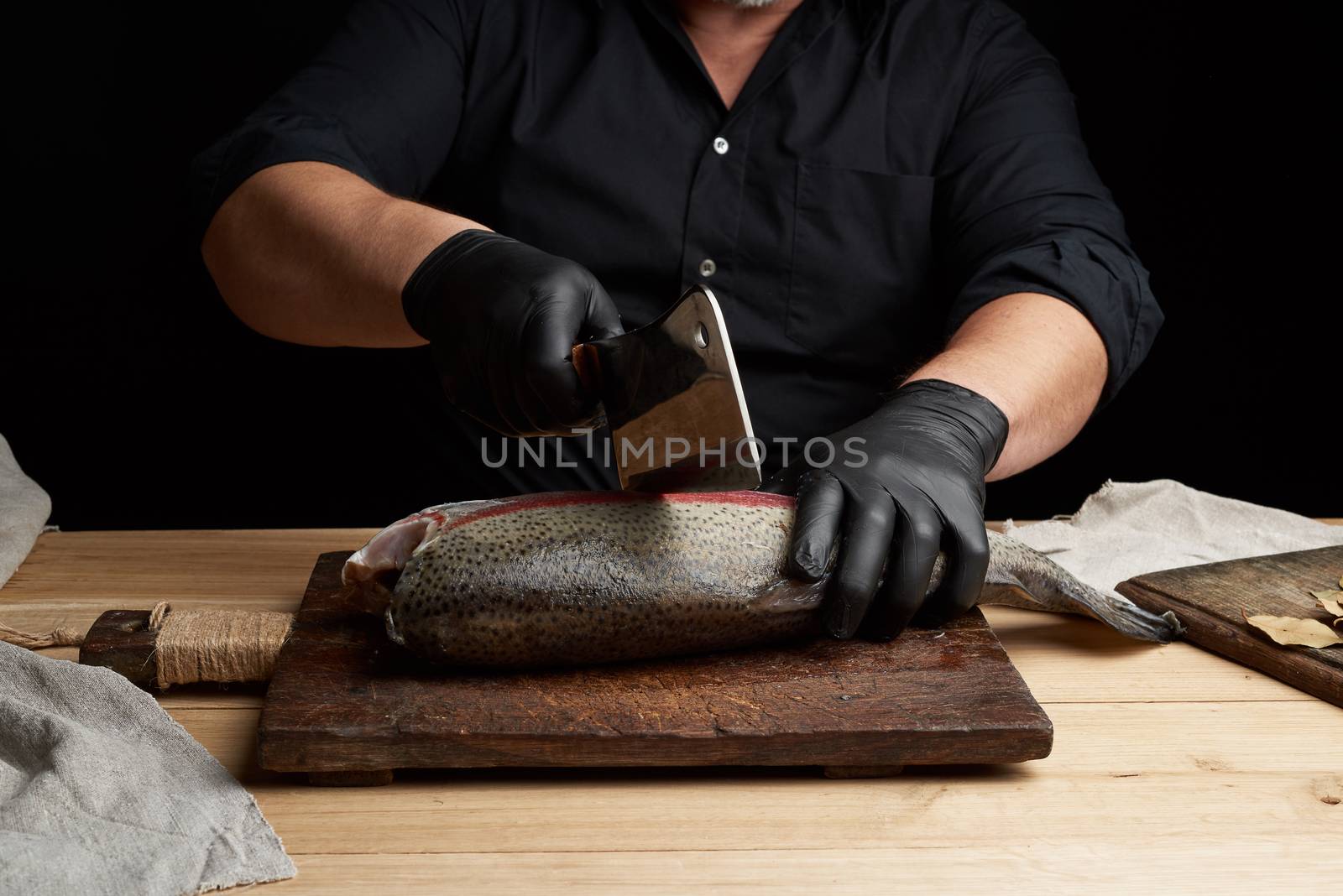 chef in black shirt and black latex gloves slices a whole fresh fish salmon on a vintage brown cutting board, dark key