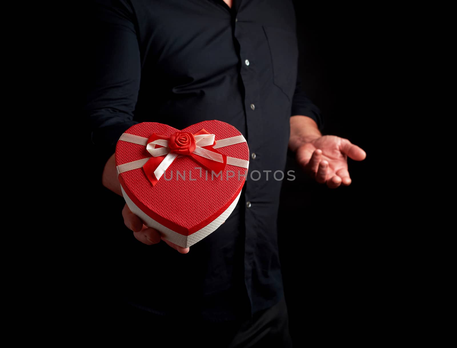 adult man in a black shirt holds a red cardboard box in the form of a heart with a bow on a dark background, concept of holiday greetings, surprise and a gift