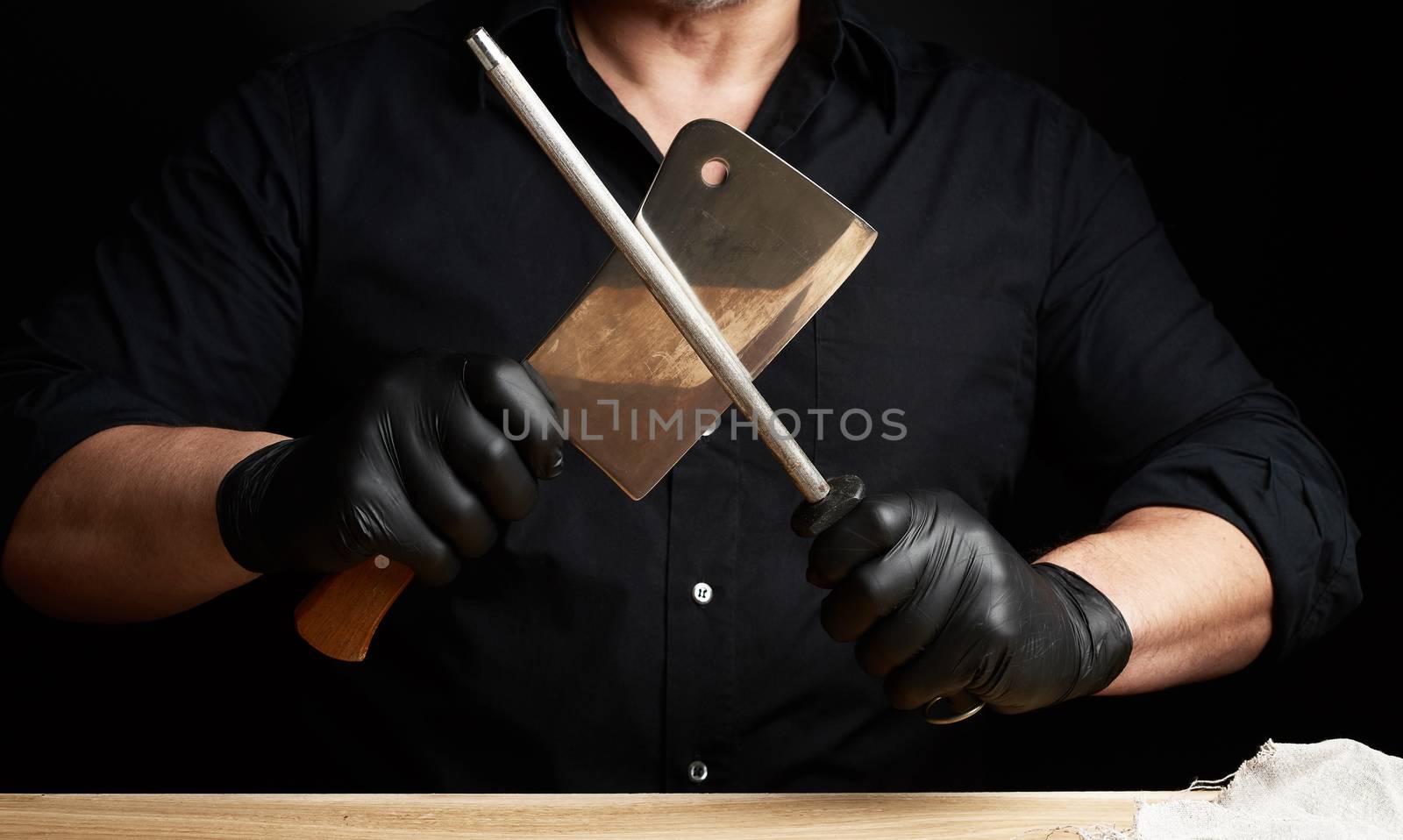 chef in a black shirt and black latex gloves sharpen a kitchen knife on an iron sharpener with a handle above the table, low key, close up