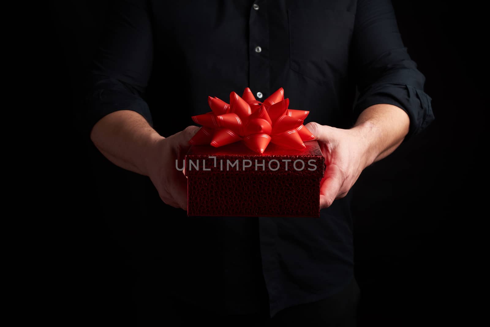adult man in a black shirt holds a red square box with a knotted bow on a dark background, concept of holiday greetings, surprise