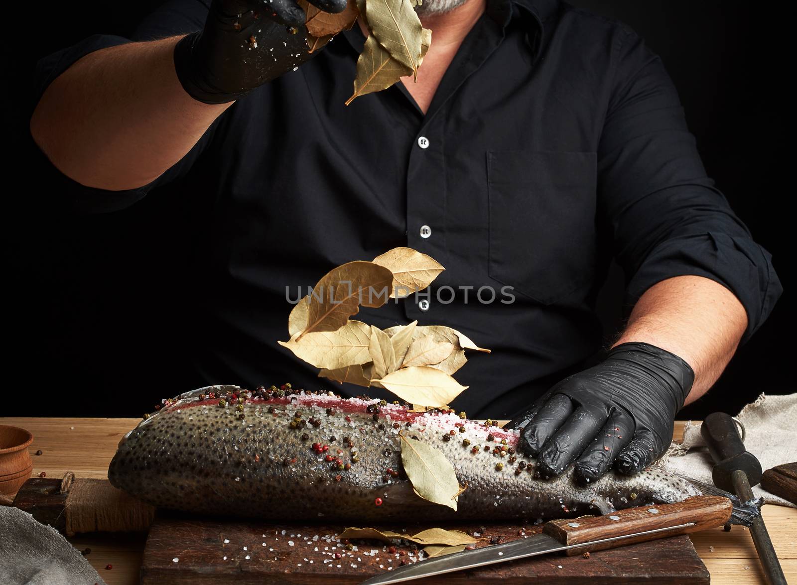 chef in black uniform and black latex gloves pours dry leaves of bay leaf onto cut fresh salmon fillet, seasoning scatters in different directions
