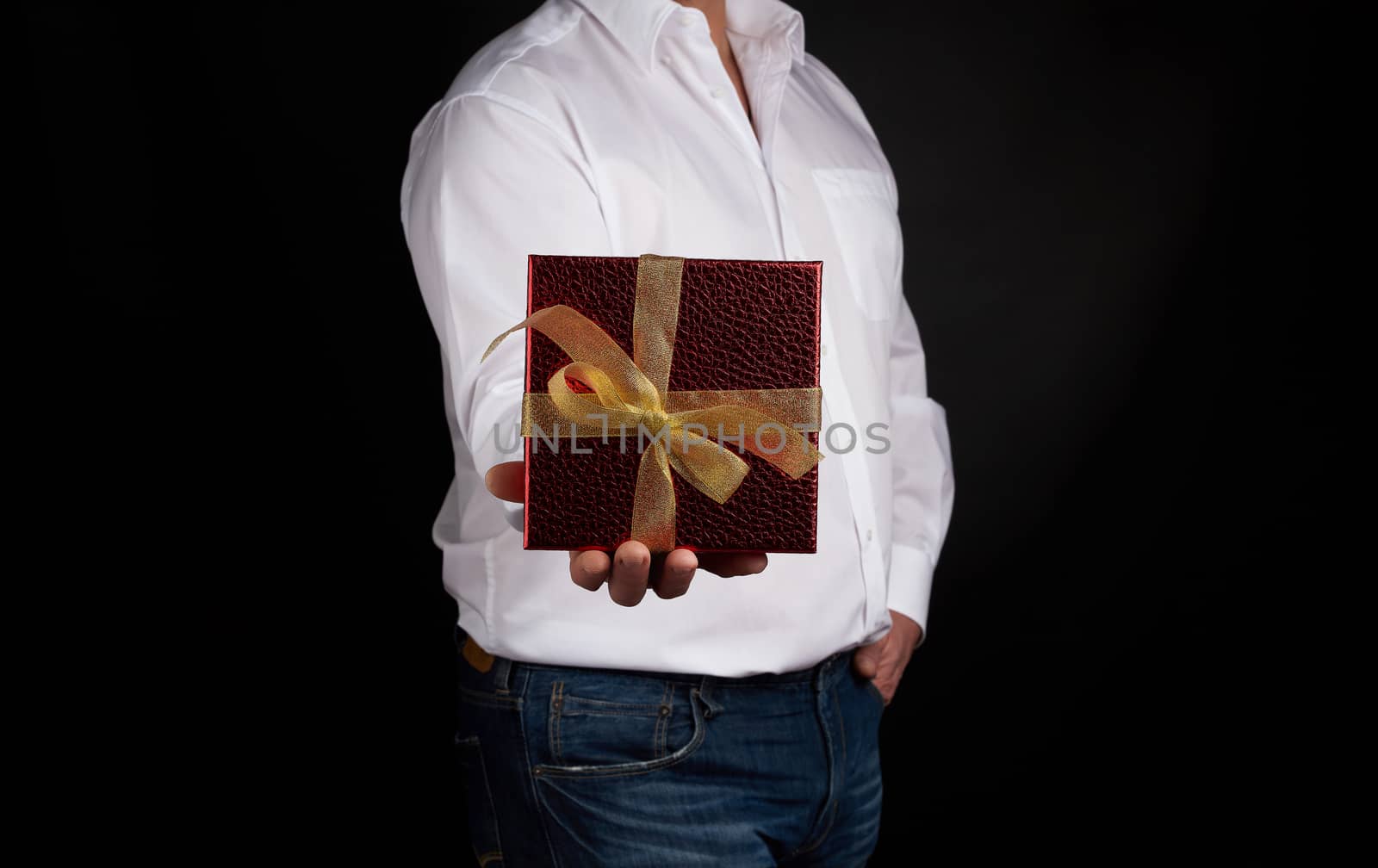 adult man in a white shirt holds a red square box with a knotted golden bow on a dark background, concept of congratulations, surprise