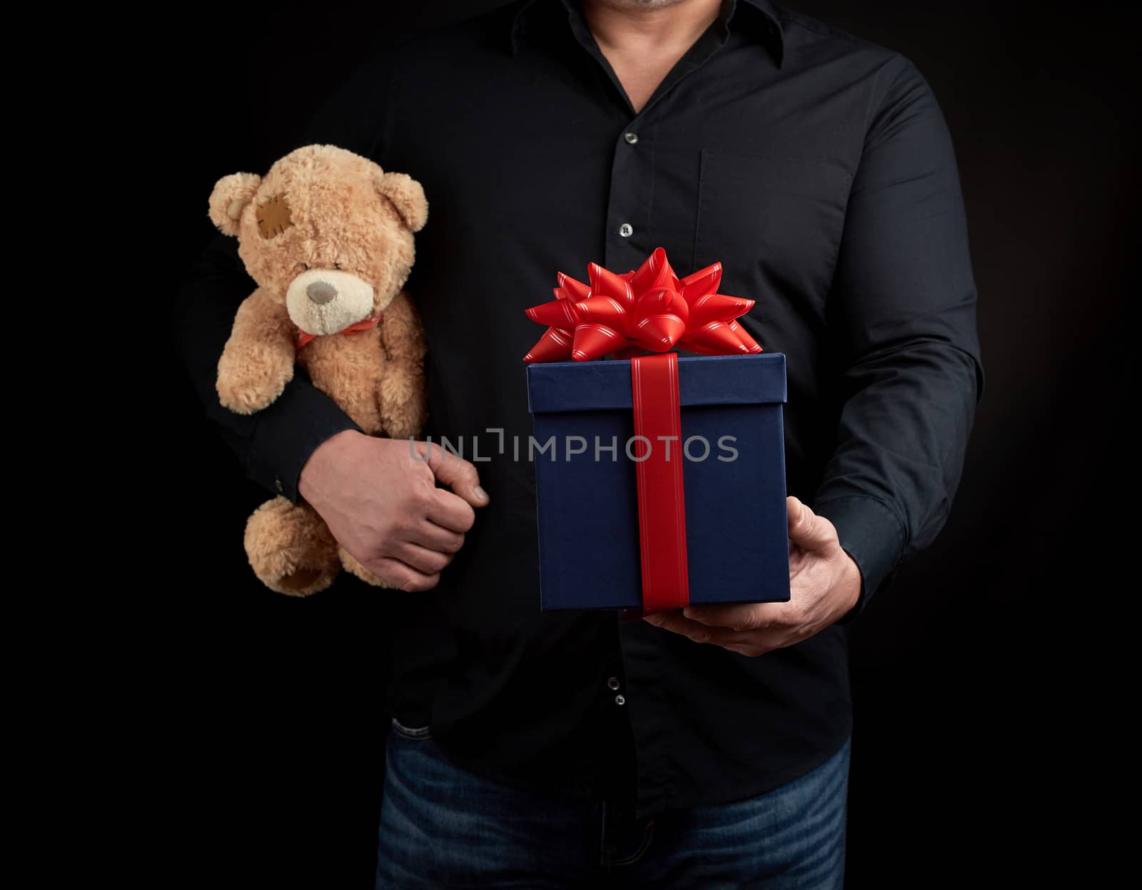 adult man in a black shirt holds a blue square box tied with a red ribbon and brown teddy bear, concept of congratulations on Valentine's Day on February 14, low key 
